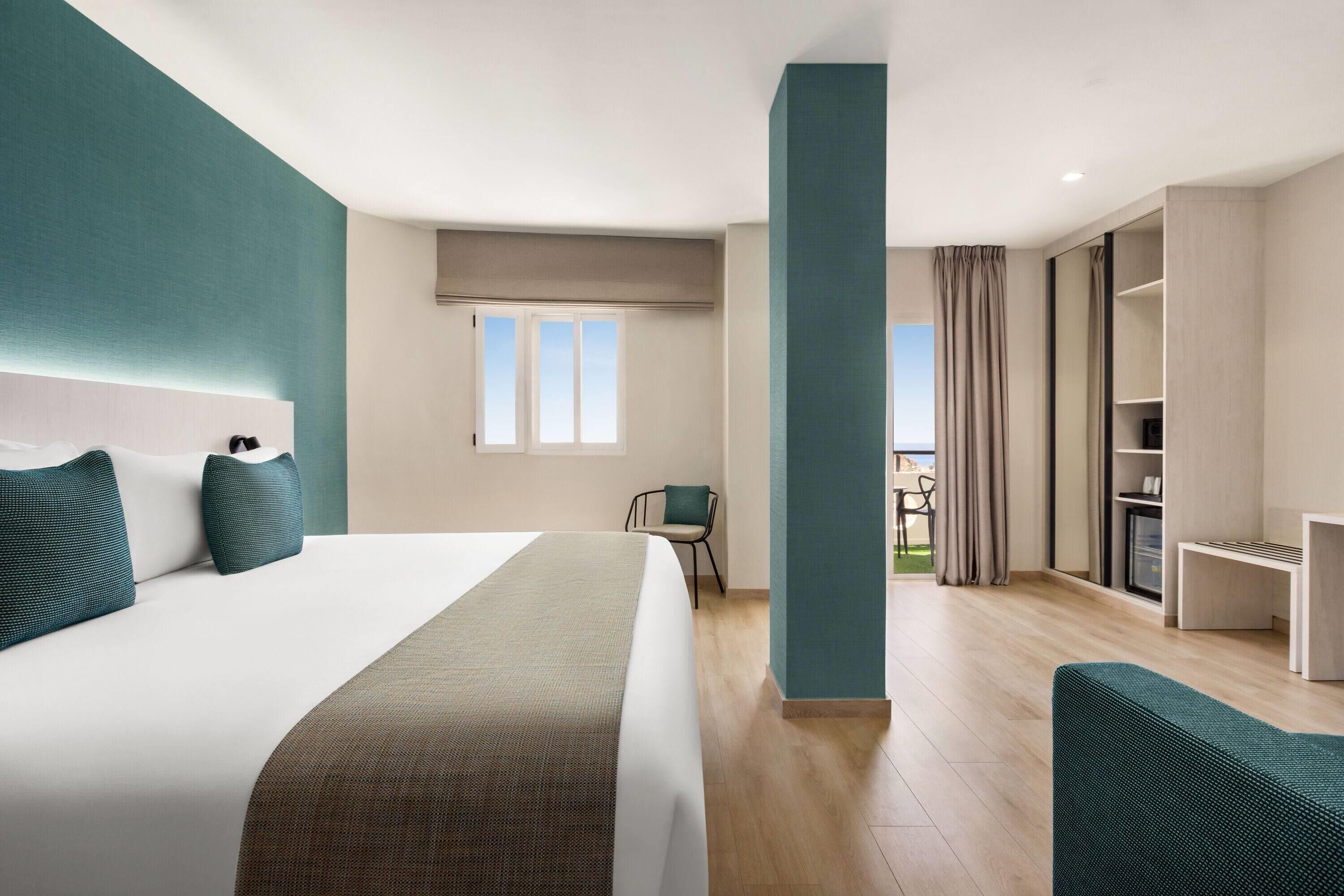 A modern hotel room with a large bed, a desk, a chair, and a view of the outdoors.