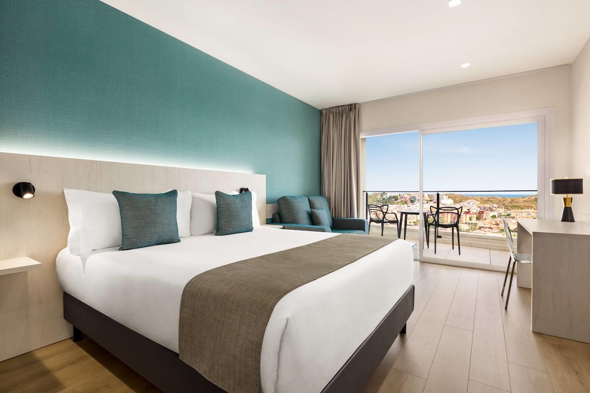 A modern hotel room with a large bed, a desk, and a balcony with a view.
