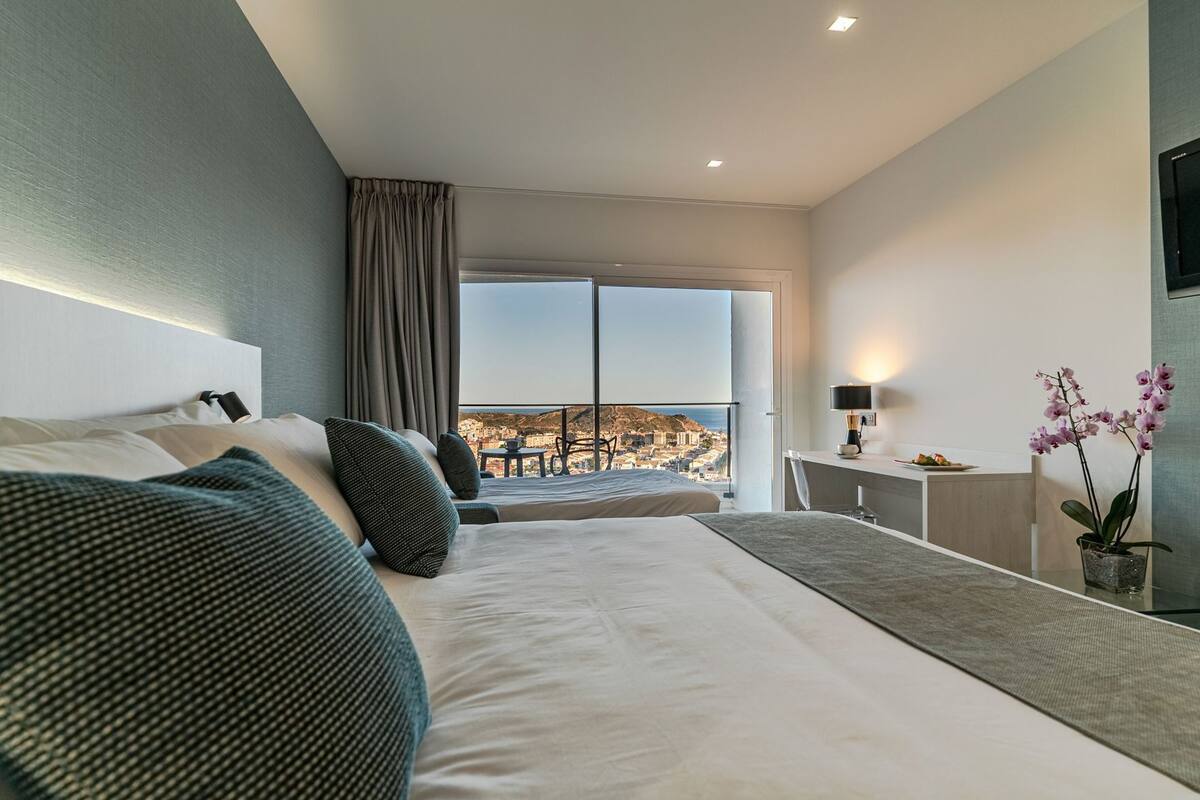 Una habitación de hotel moderna con una cama grande, un escritorio, una silla, una orquídea en maceta y vistas a un paisaje urbano a través de un ventanal amplio.