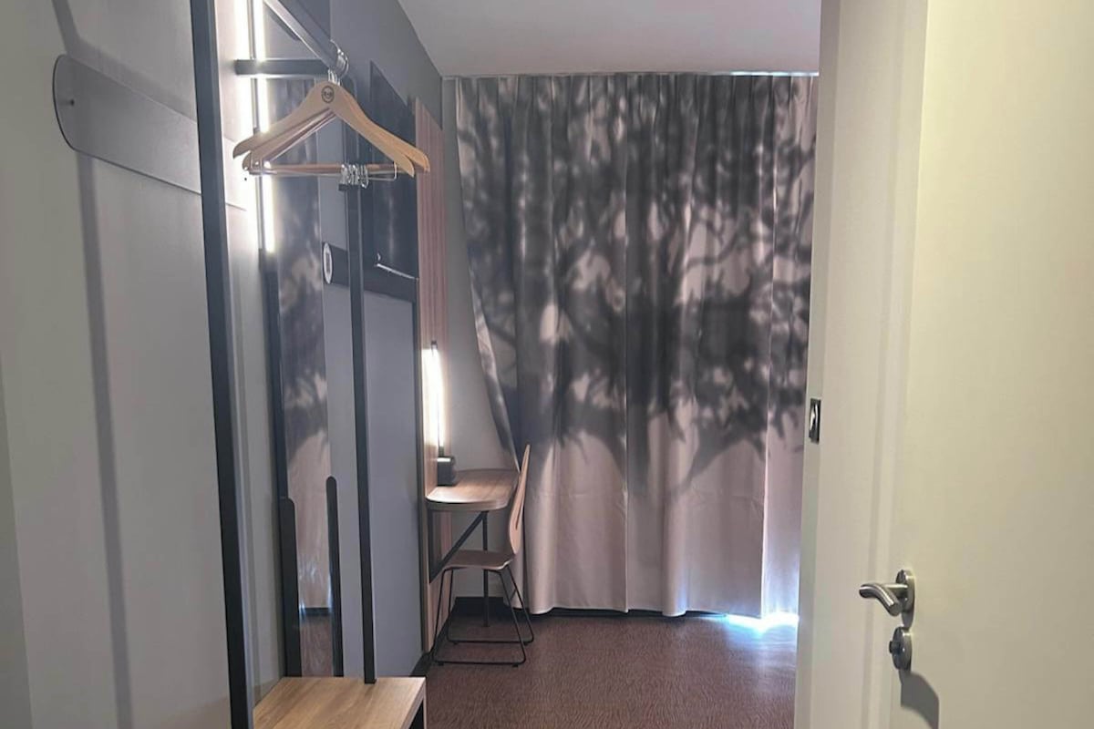 A hotel room with a wooden floor, a chair, a table, a hanging coat rack, and a curtain with a tree pattern.