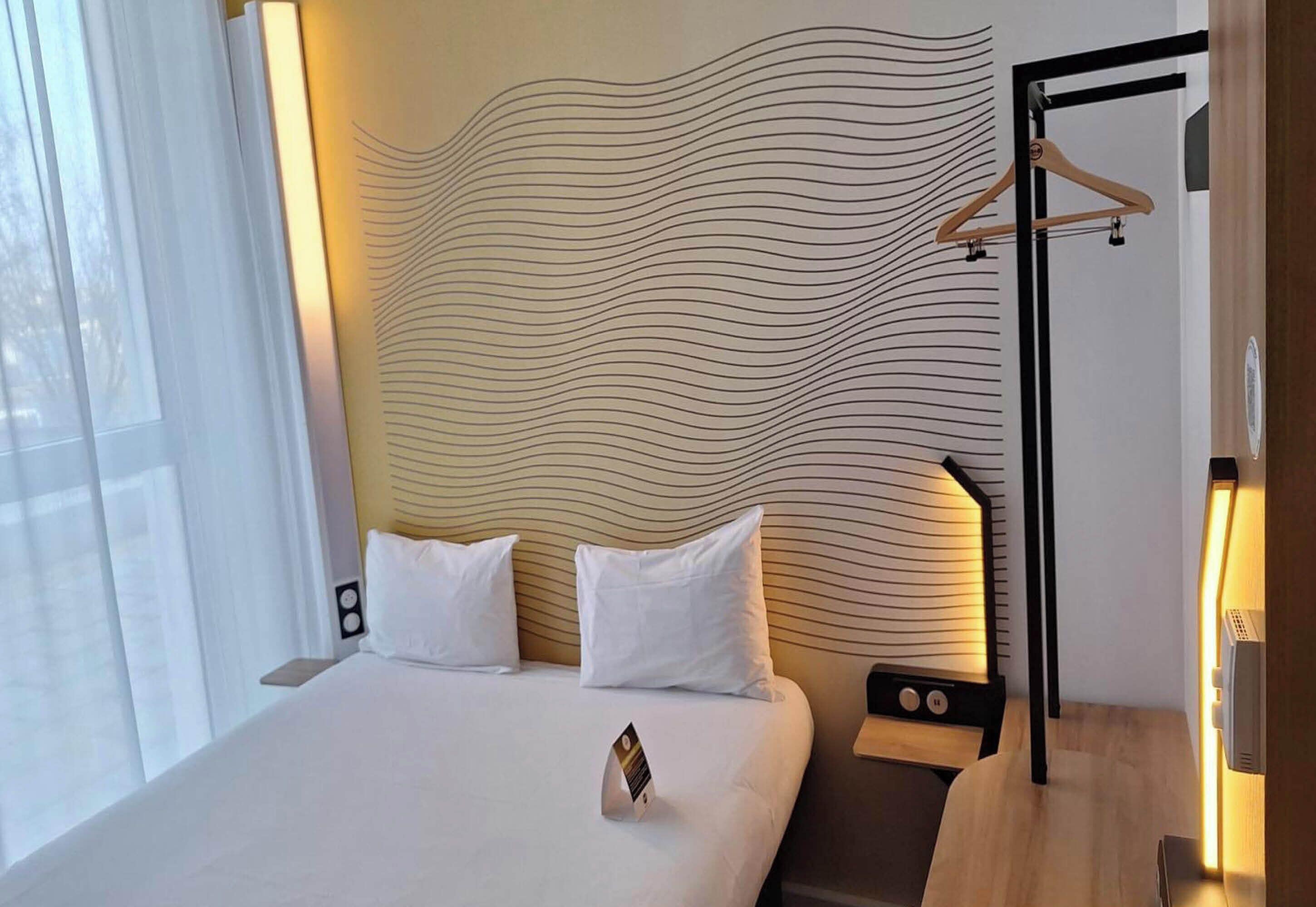 A hotel room with a bed, two pillows, a bedside table with a lamp, and a wall with a wavy pattern.
