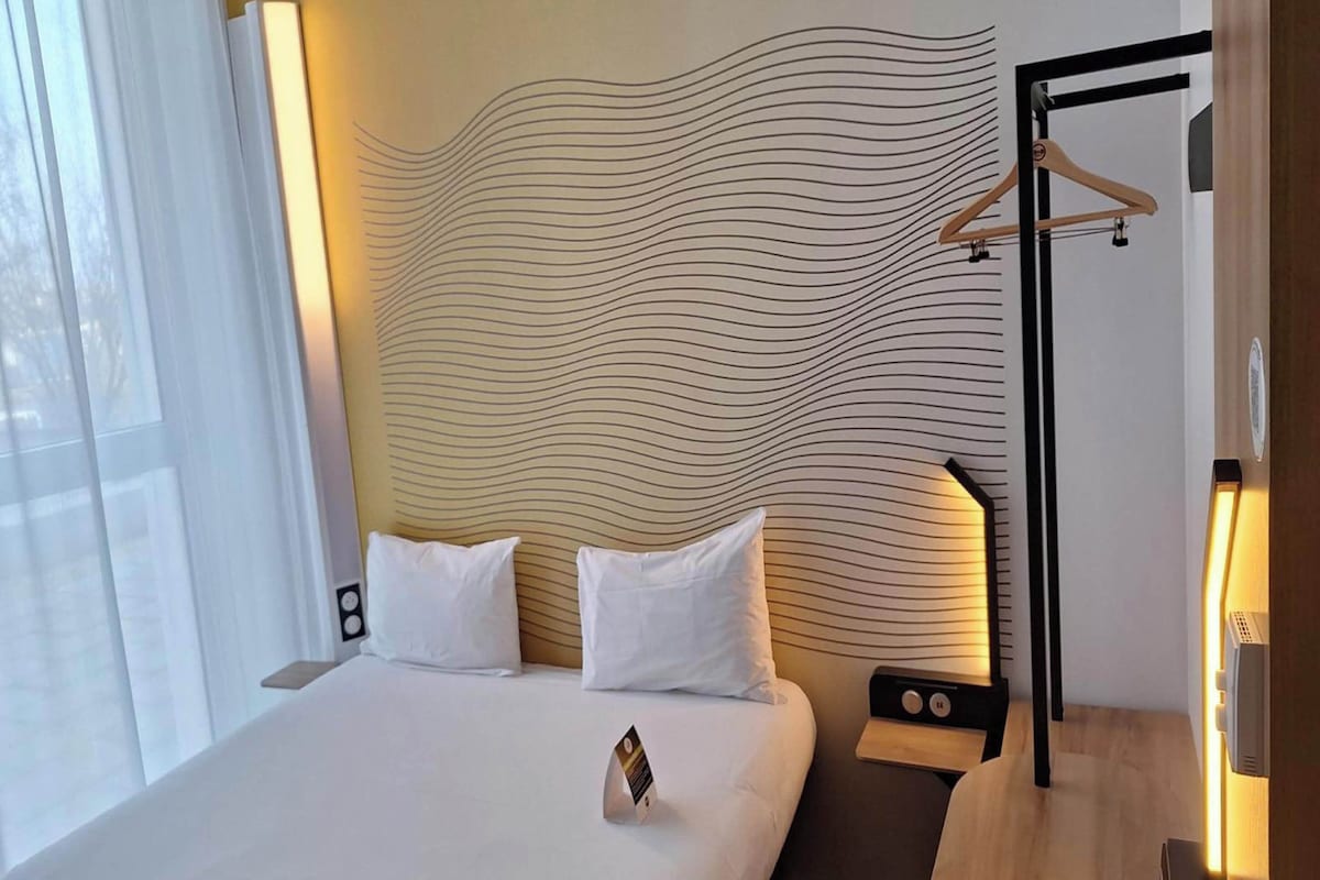 A hotel room with a bed, two pillows, a bedside table with a lamp, and a wall with a wavy pattern.