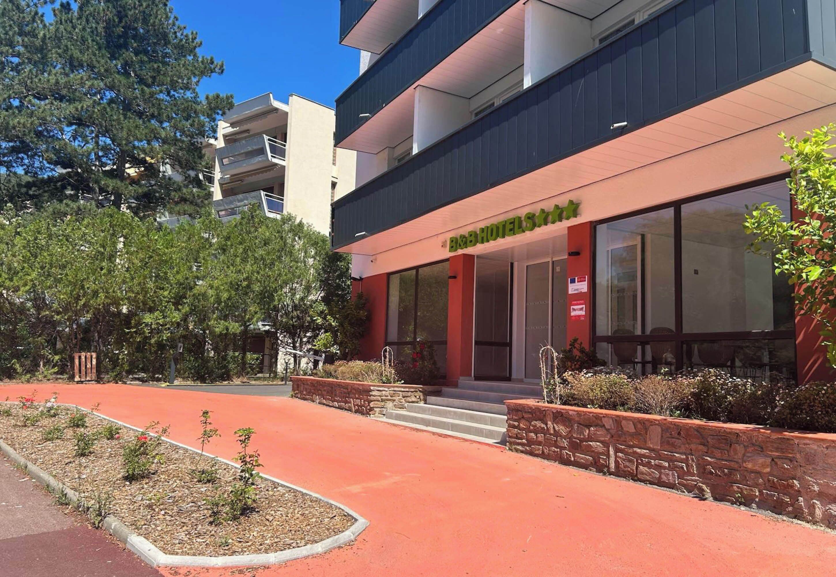 A hotel with a modern facade, a red-paved entrance, and a landscaped garden area.