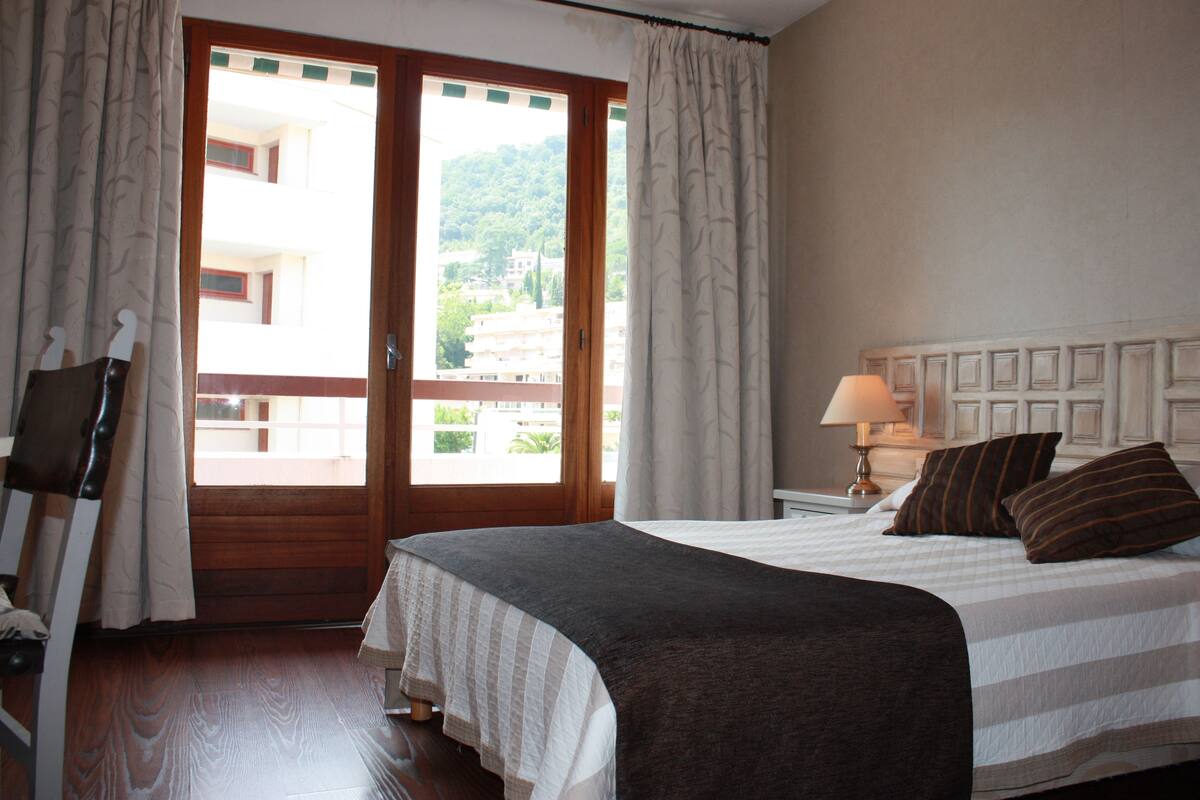 A hotel room with a bed, a nightstand, a lamp, and a view of buildings through a window.