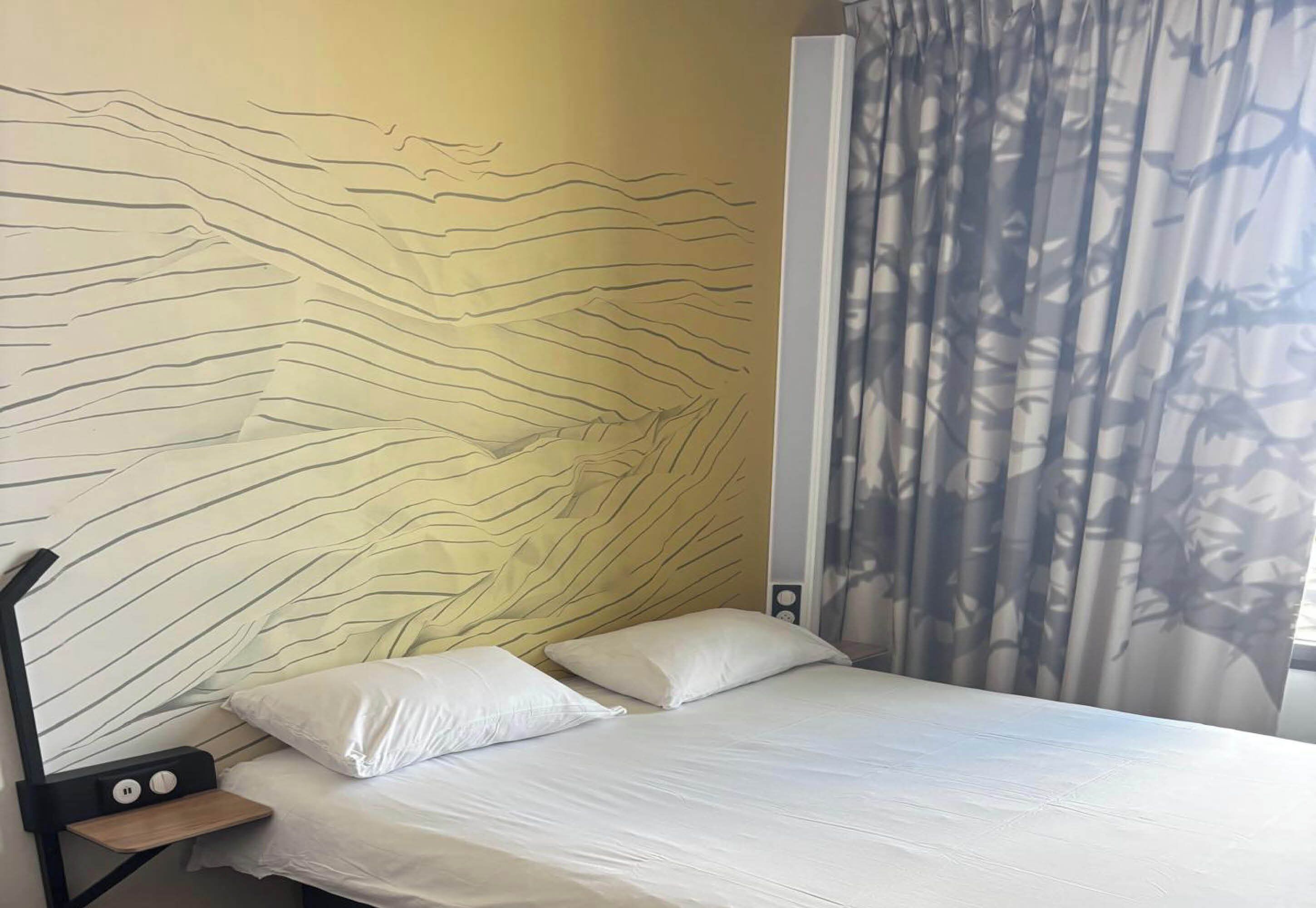 A bed with white bedding, a headboard with a wavy design, and a window with a curtain.