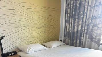 A bed with white bedding, a headboard with a wavy design, and a window with a curtain.