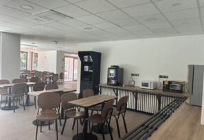 A cafeteria with tables and chairs, a coffee machine, a microwave, and a refrigerator. - B&B HOTEL Amélie-les-Bains Centre (Amelie-les-Bains-Palalda)