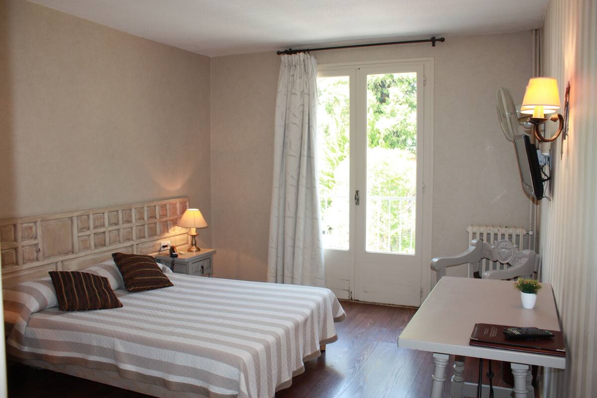 A hotel room with a bed, bedside table, lamp, and a small table with a plant and remote control.
