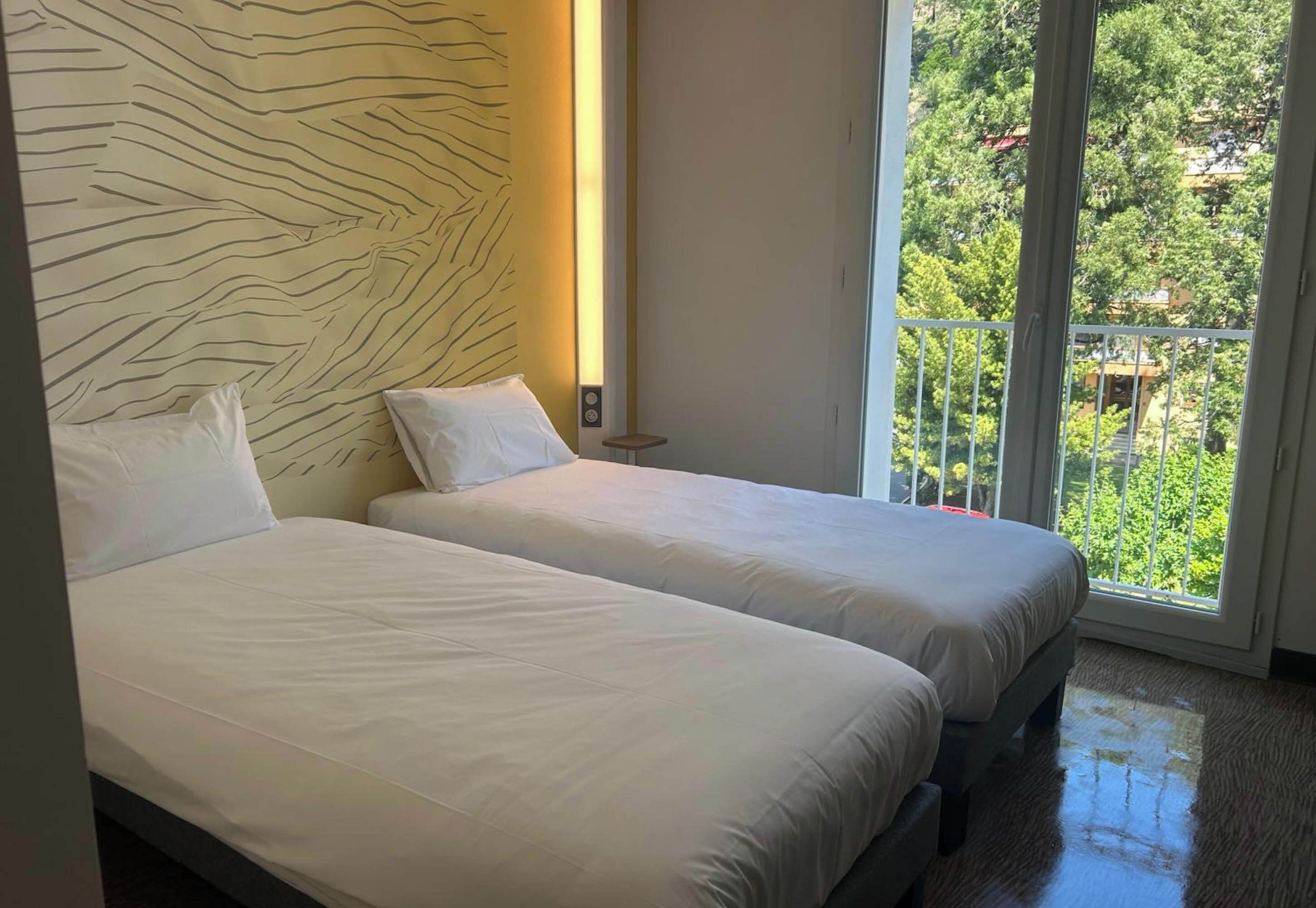 Une chambre d’hôtel avec deux lits, une grande fenêtre et une vue sur la verdure environnante.