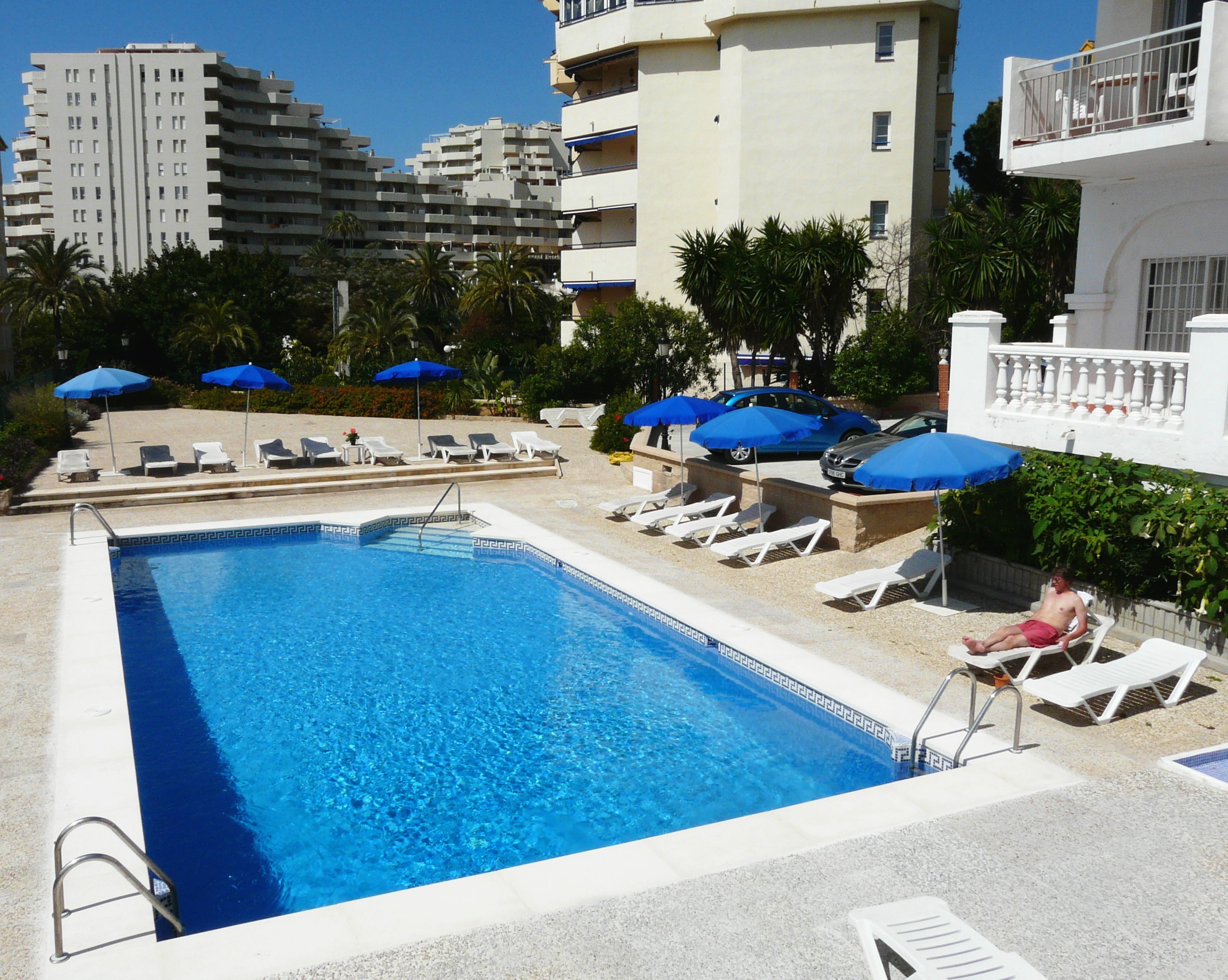 Piscina con sillas de descanso y sombrillas, rodeada de edificios.