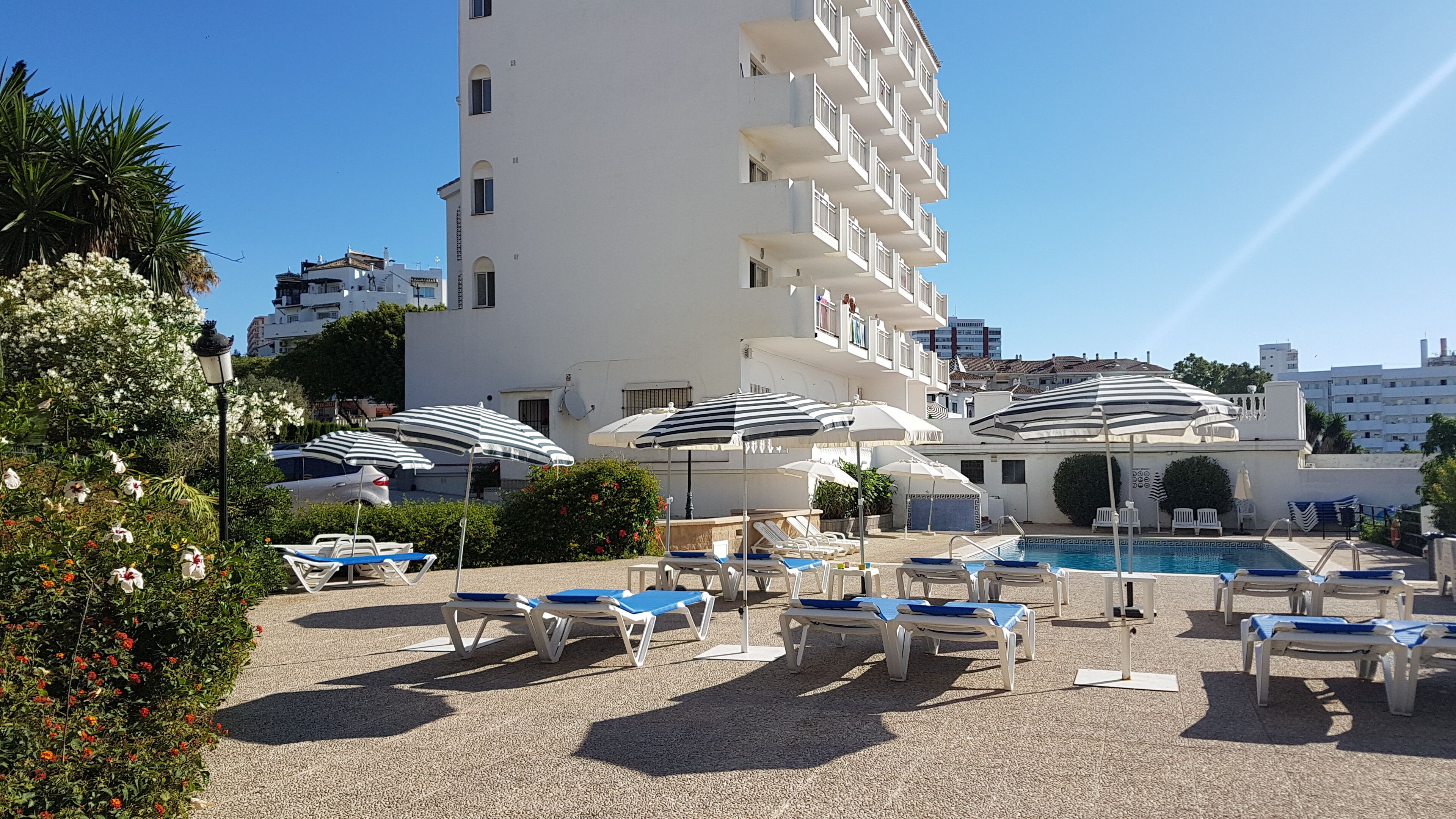 Un espace aménagé au bord de la piscine, comprenant plusieurs chaises longues et des parasols, ainsi qu’une piscine et un bâtiment blanc doté de balcons.