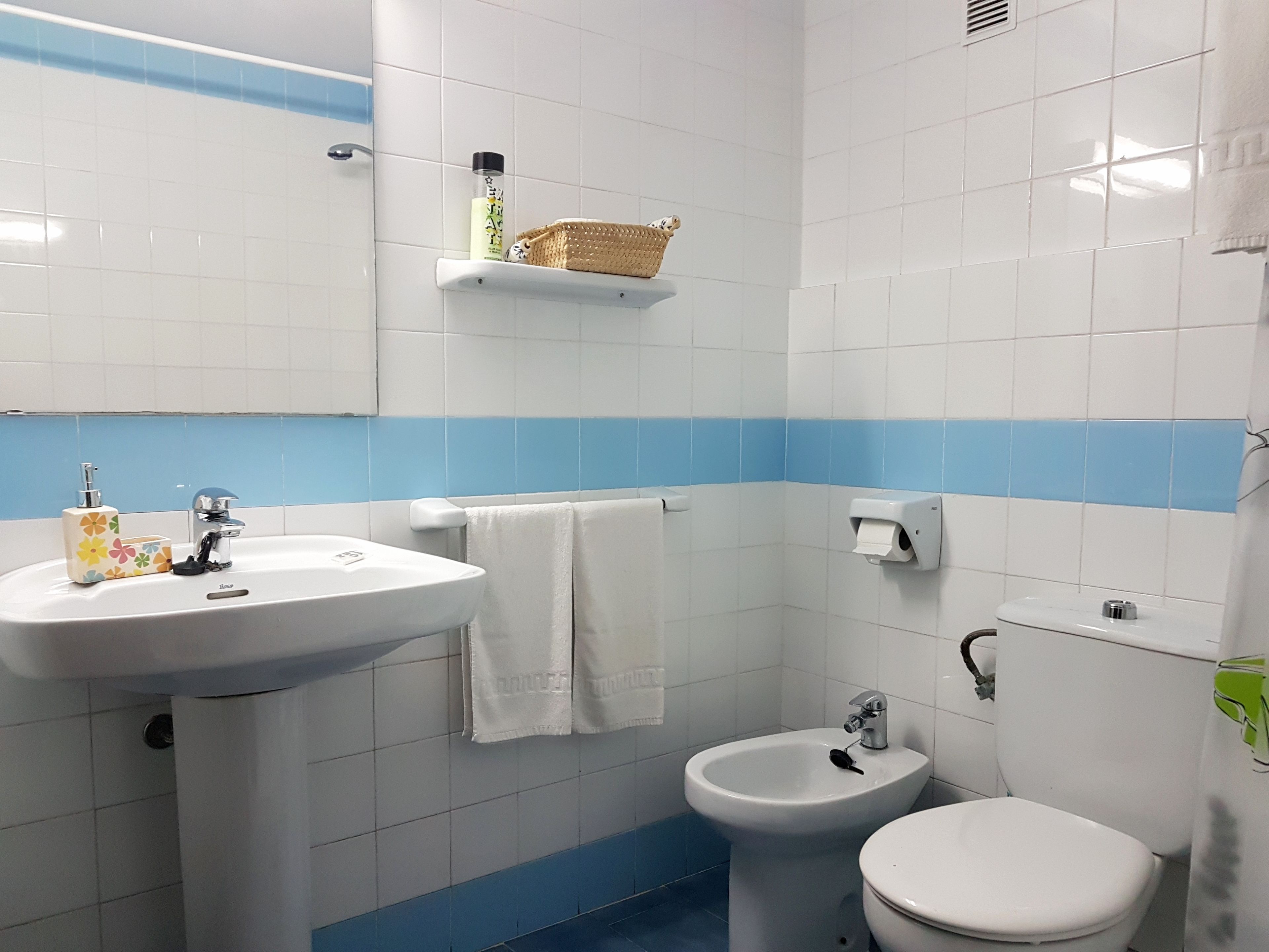 Un baño con lavamanos, bidé e inodoro. Hay un perchero, dispensador de jabón y una cesta en la pared.