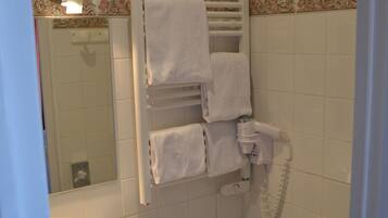 Un baño con lavamanos, inodoro y espejo. Hay toallas colgadas en un perchero y se ve un cabezal de ducha.