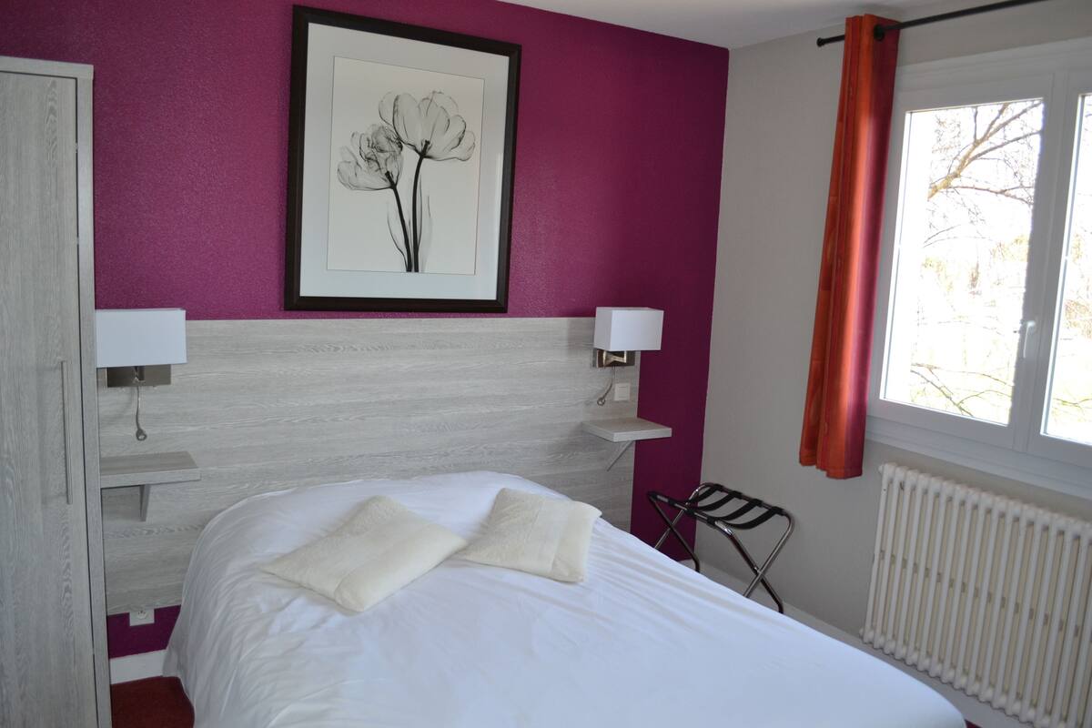 Un dormitori amb un llit, dues taules de nit amb làmpades, una obra d'art emmarcada a la paret, una finestra amb cortines i un radiador.
