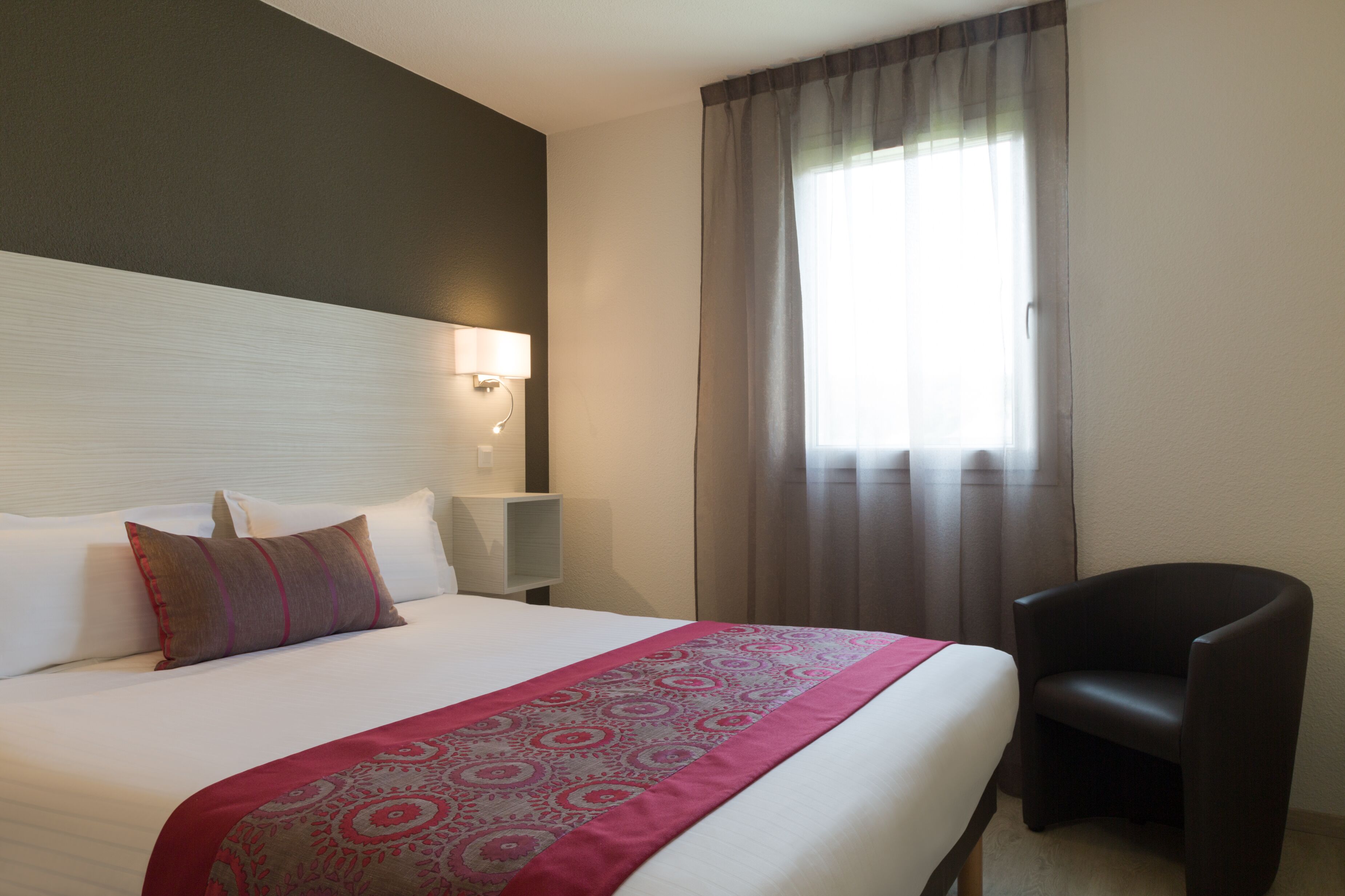 A hotel room with a bed, a chair, a nightstand, and a window with curtains.