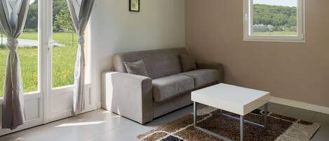 A modern living room with a sofa, a coffee table, and a view of a green landscape through a window.