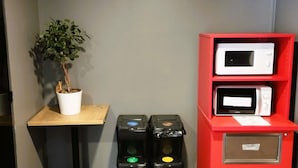 A room with a potted plant on a small table, a red shelf with two microwaves, and two trash bins with recycling symbols. - hotelF1 Lyon Bourgoin-Jallieu (L'Isle-d'Abeau)
