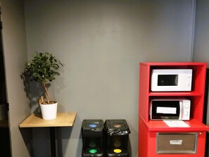 Una habitación con una planta en maceta sobre una mesita, un estante rojo con dos microondas y dos botes de basura con símbolos de reciclaje.