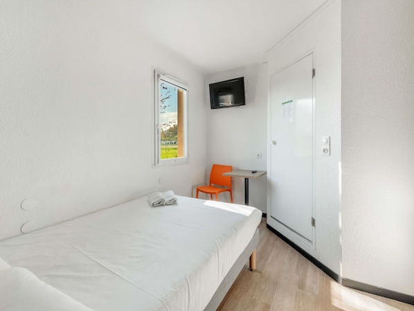 A small, single-bed room with a window, a chair, a small table, and a TV mounted on the wall.