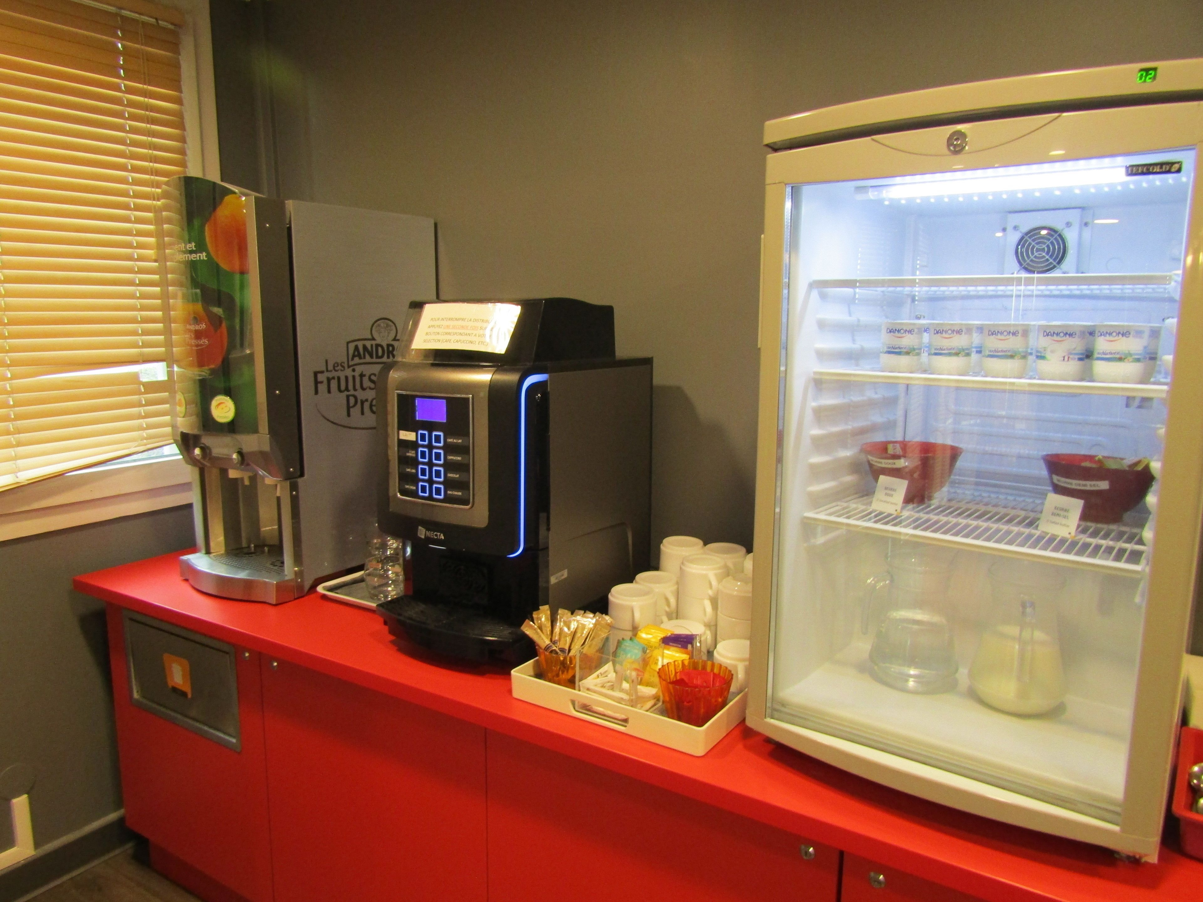 A self-service coffee station with a digital coffee machine, a refrigerator stocked with beverages, and a display of various coffee pods and accessories.