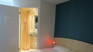 Habitación de hotel con cama, mesita de noche con luz roja, baño con lavamanos y espejo, y puerta que da a otra habitación.