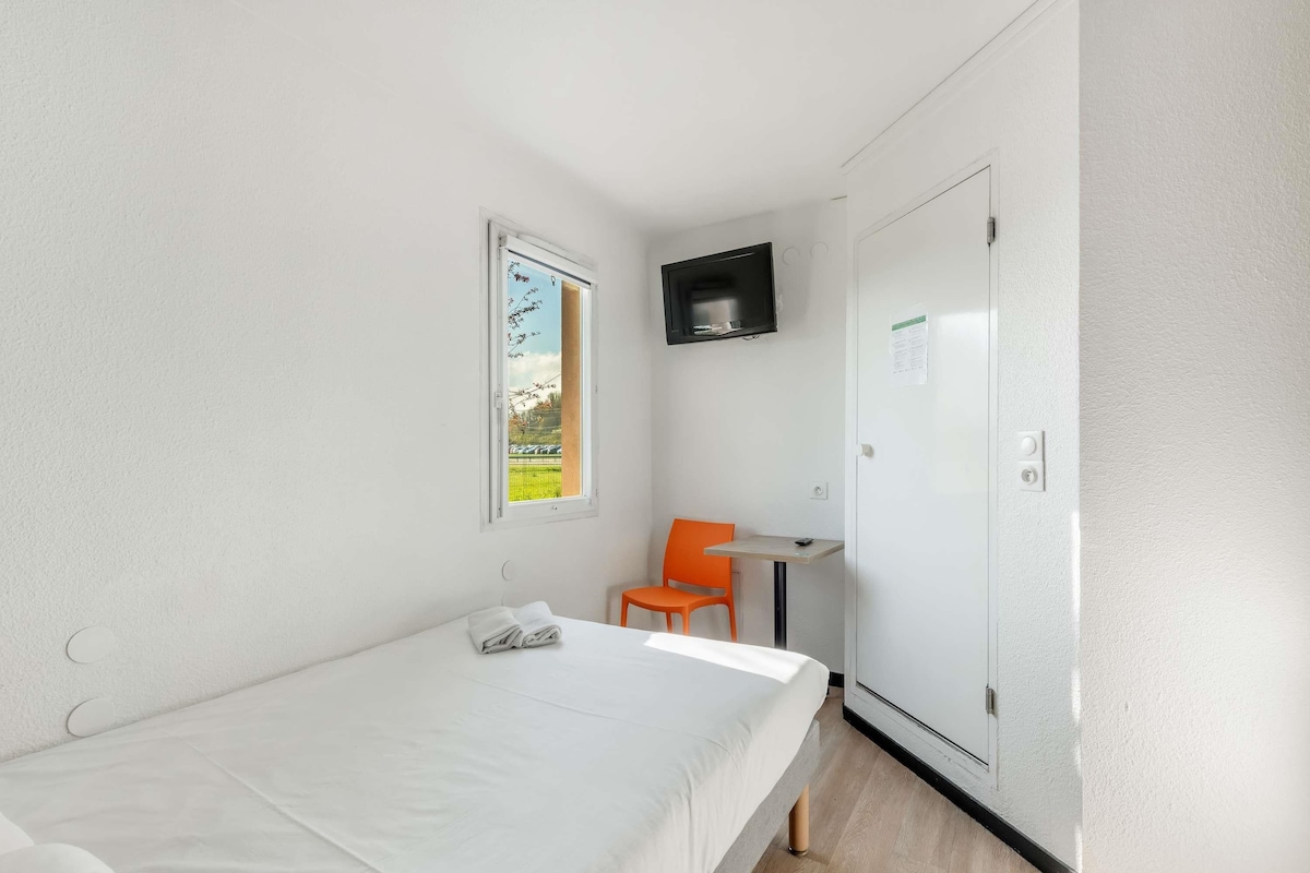 Una habitación pequeña con cama individual, ventana, una silla, una mesita y televisor montado en la pared.