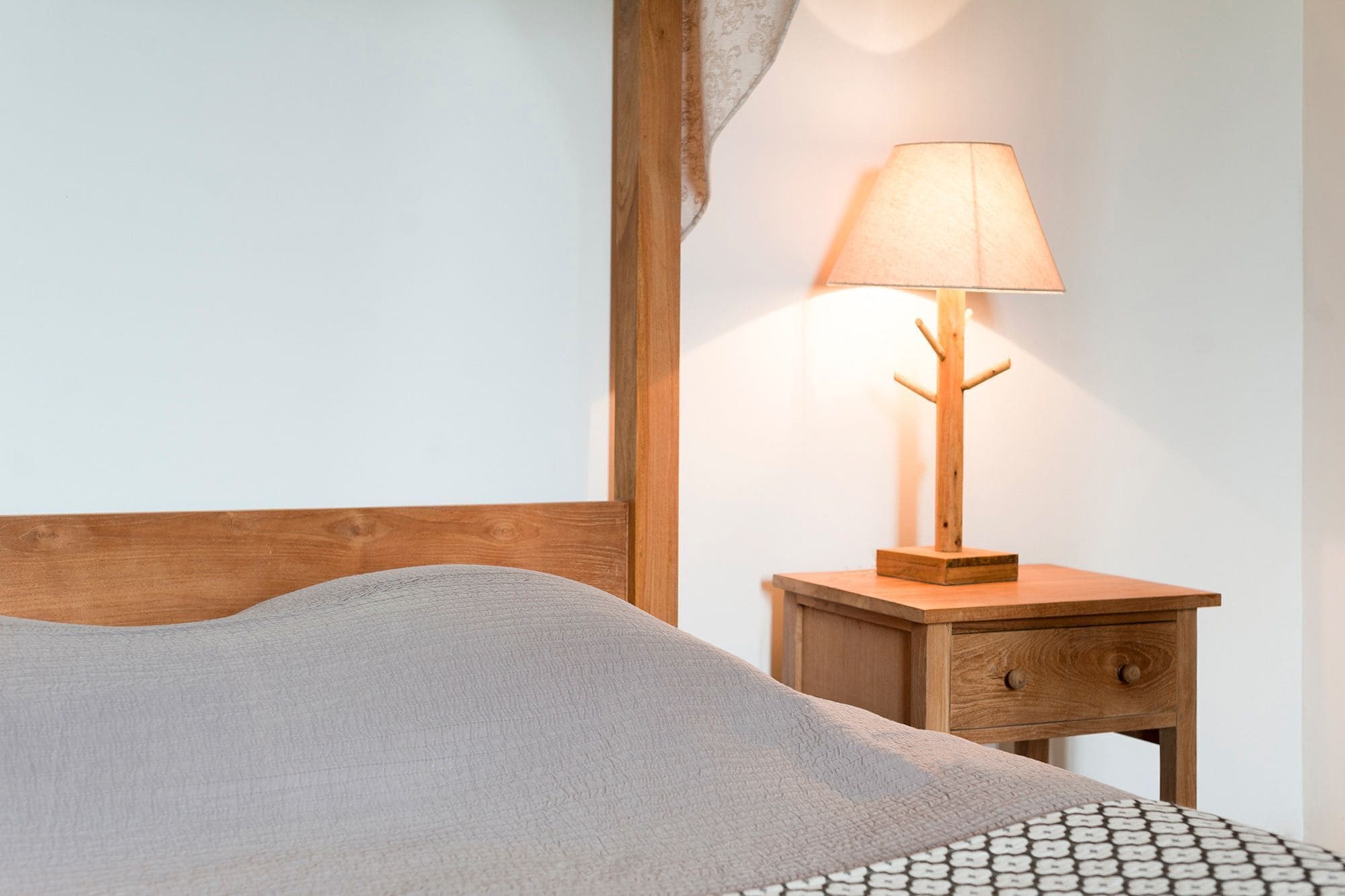Una cama bien hecha con una manta gris, una mesita de noche de madera con una lámpara y un cajón pequeño.