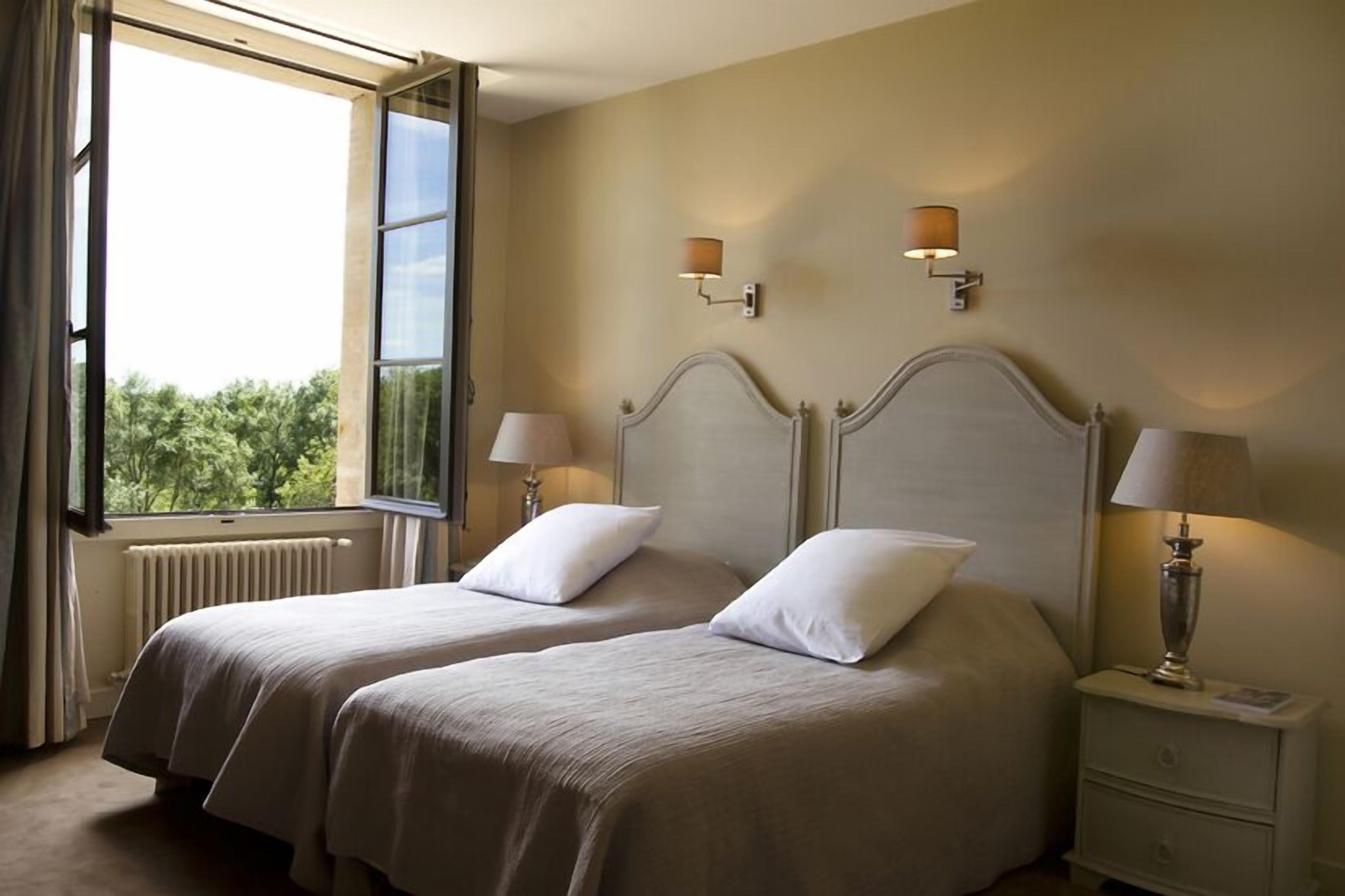 A hotel room with two beds, a window with curtains, a radiator, and two wall-mounted lamps.