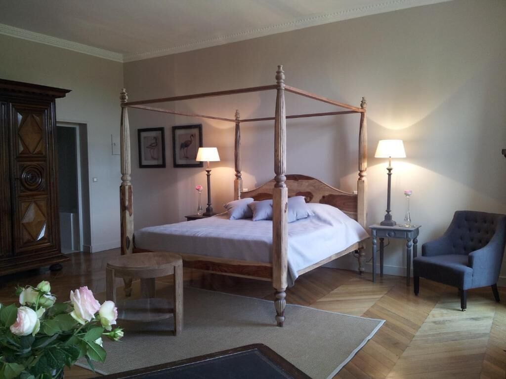A four-poster bed with a wooden frame, a bedside table with a lamp, a chair, and a small table with flowers.