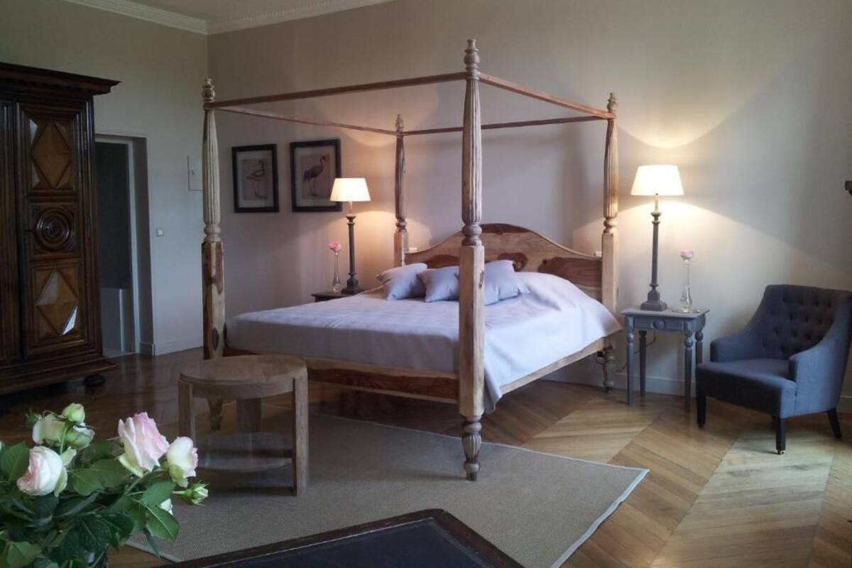 A four-poster bed with a wooden frame, a small wooden stool, a chair, and a nightstand with a lamp.