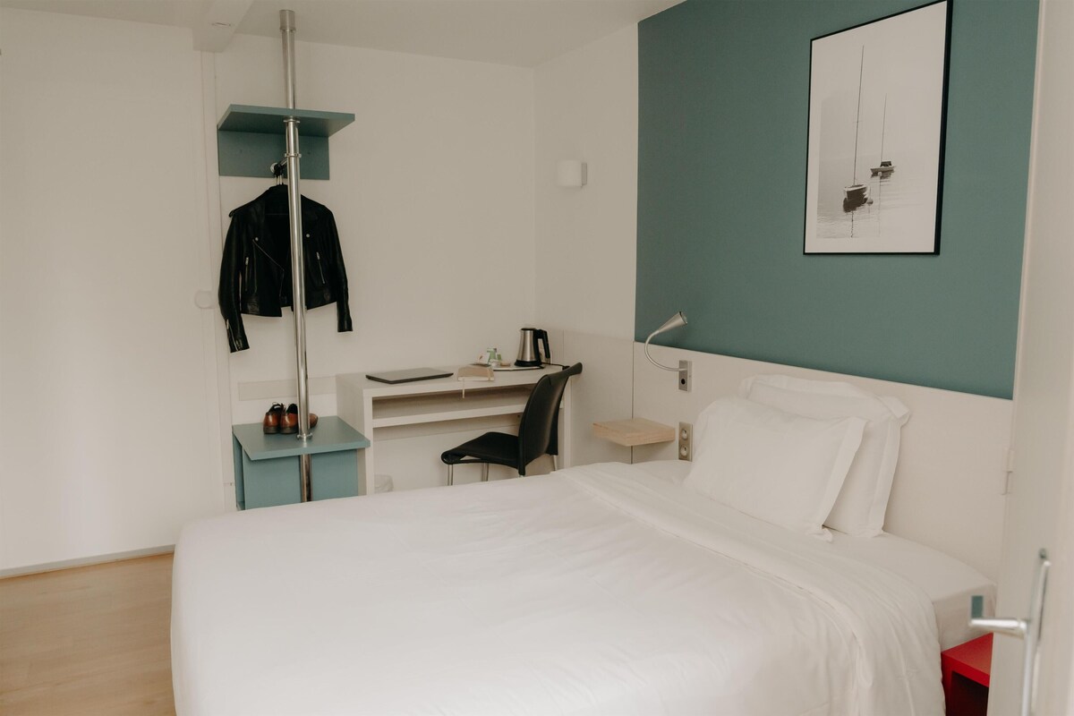 A hotel room with a bed, a desk, a chair, a coat rack, and a framed picture on the wall.
