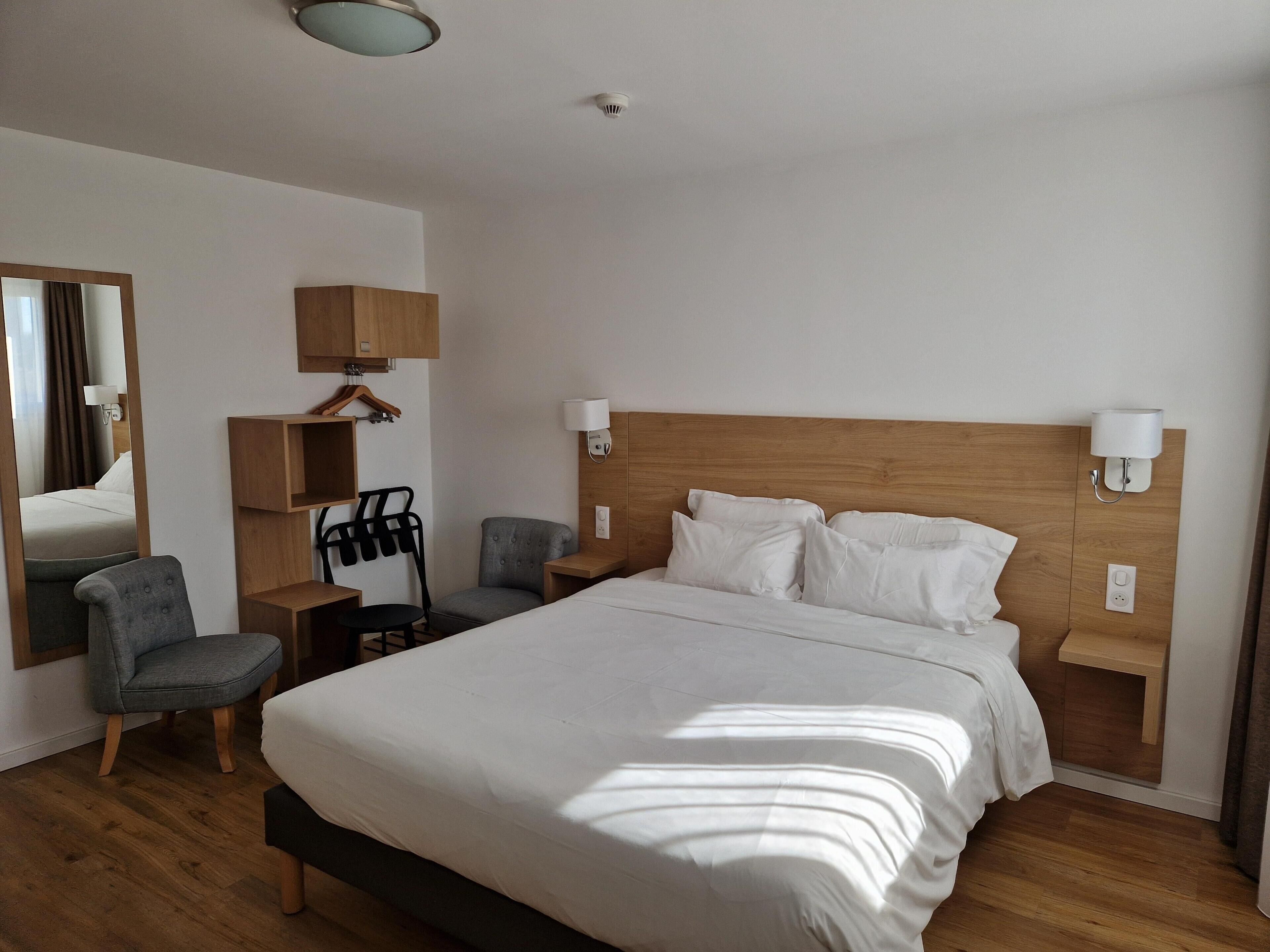 A hotel room with a large bed, a chair, a desk, a mirror, and a lamp.