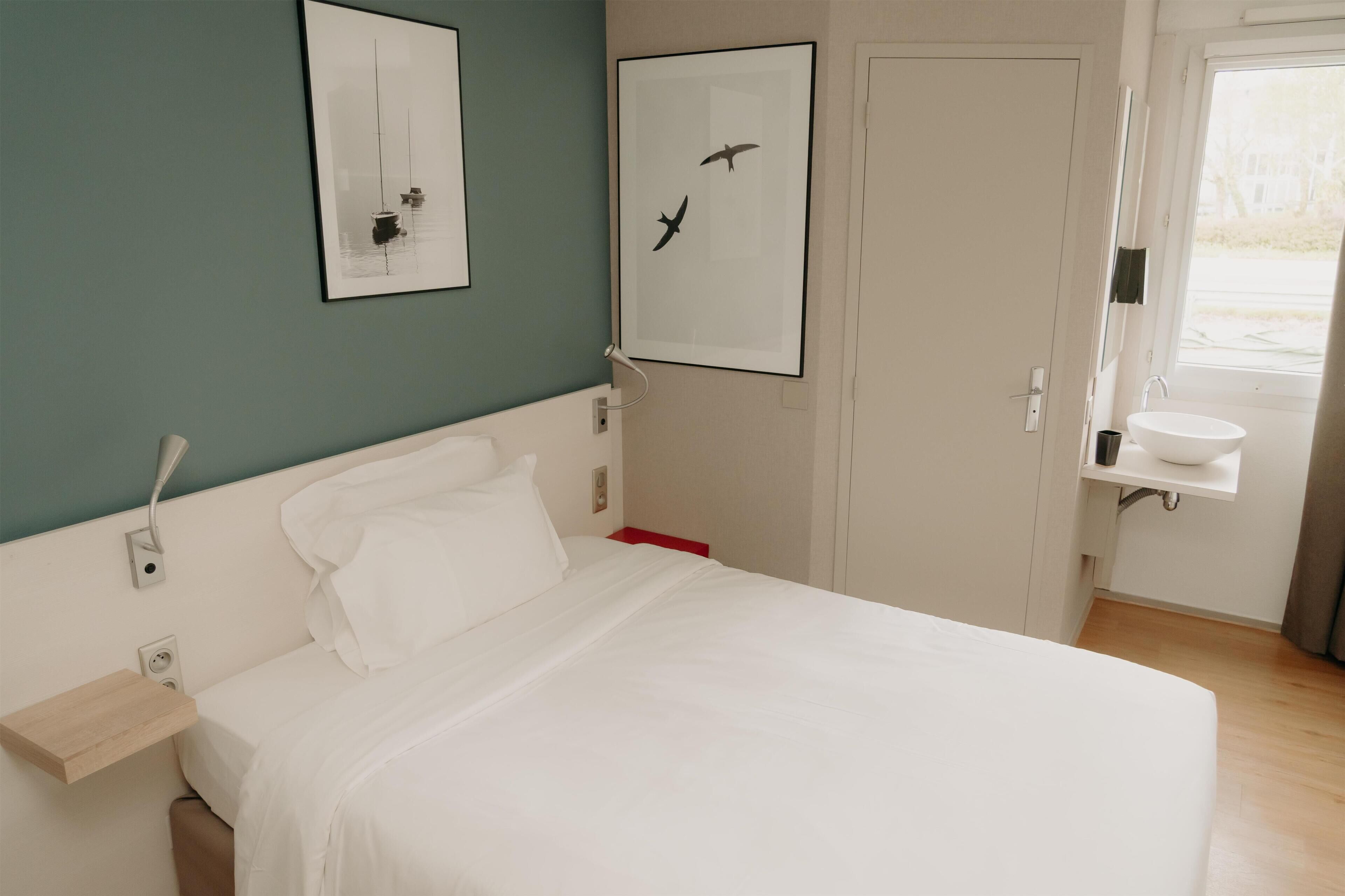 A bedroom with a bed, a bedside table, a wall-mounted light, a framed picture, a door, and a window.