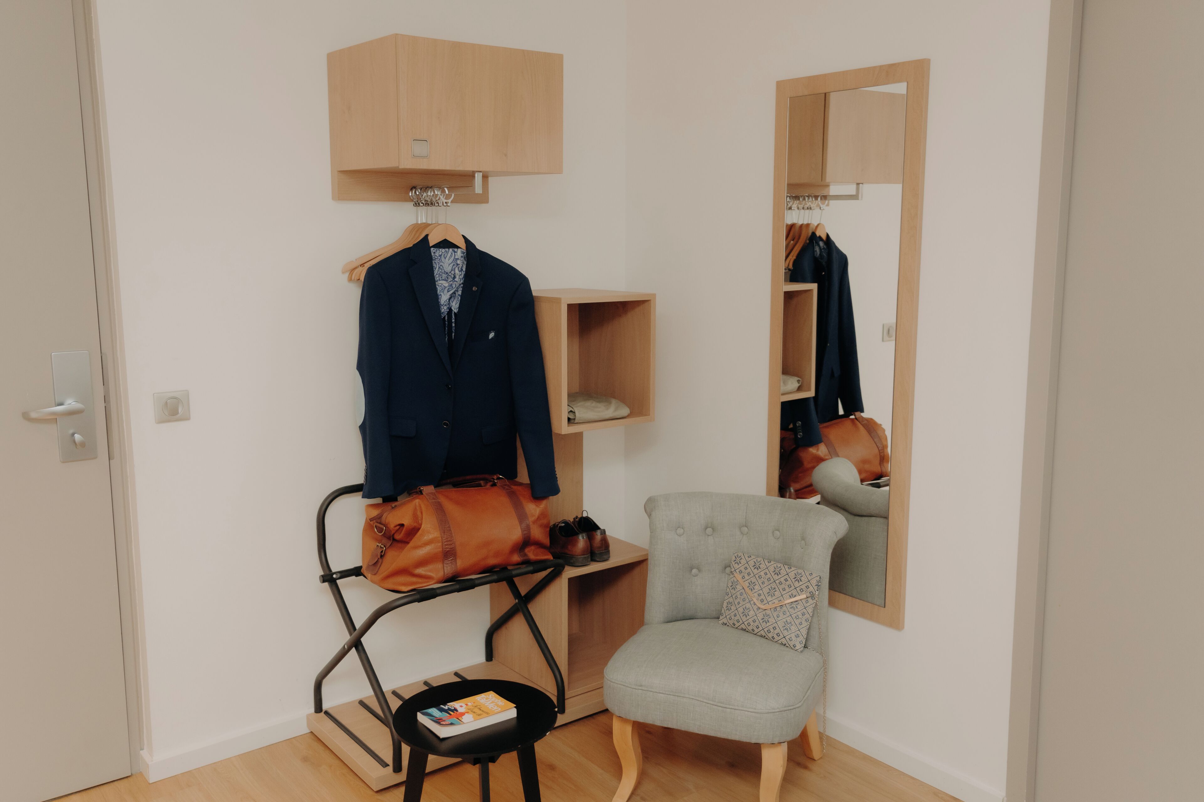 A room with a wooden wardrobe, a chair, a small table with a book, and a hanging bag.