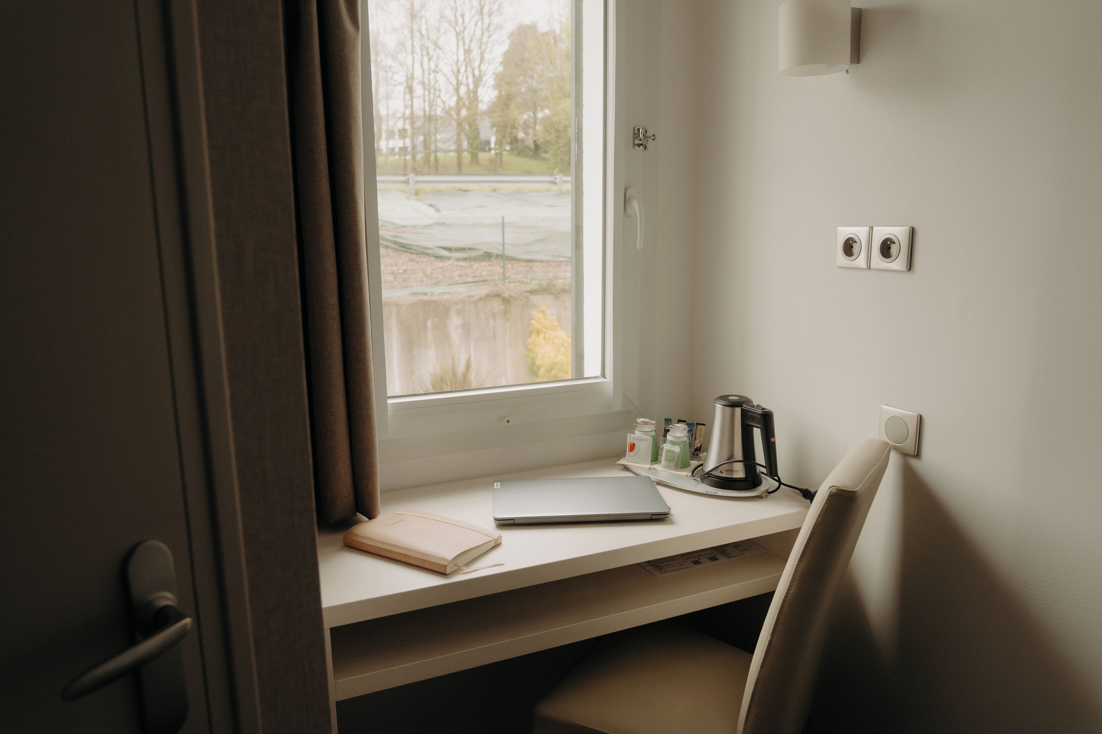 A room with a desk, a laptop, a notebook, a kettle, and a window with a view of trees and a fence.