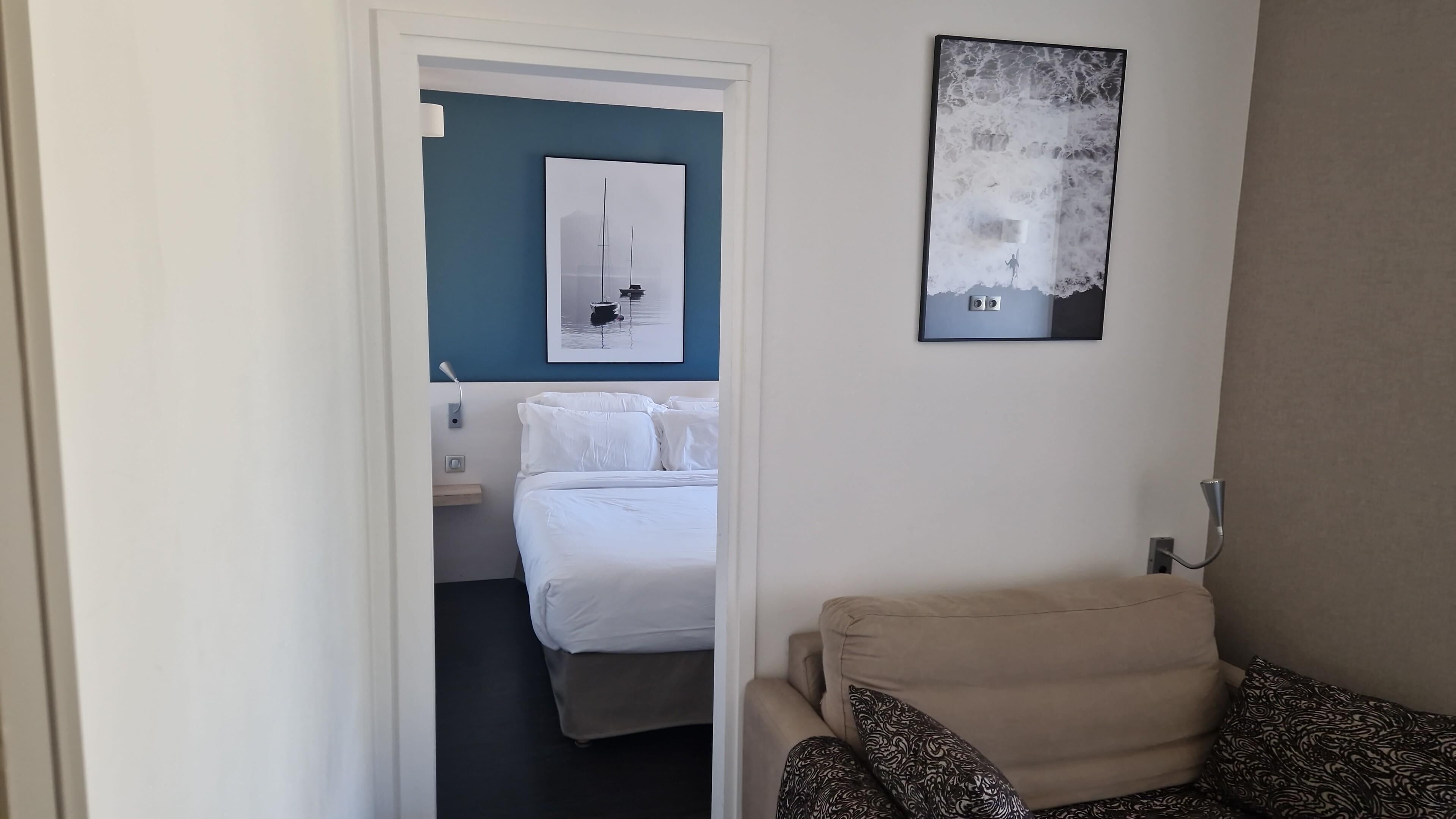 A hotel room with a bed, a chair, a wall-mounted artwork, and a door leading to another room.