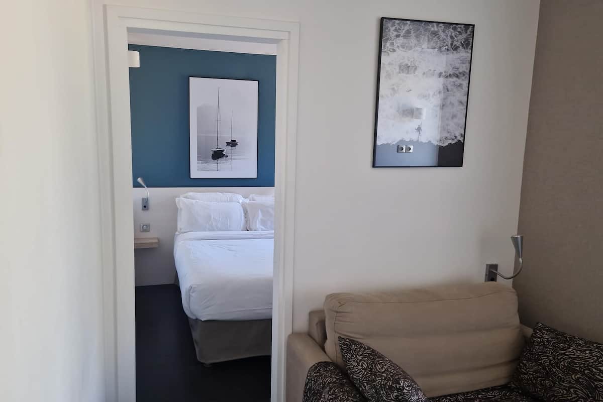 Una habitación de hotel con una cama, una silla, una obra de arte colgada en la pared y una puerta que da a otra habitación.