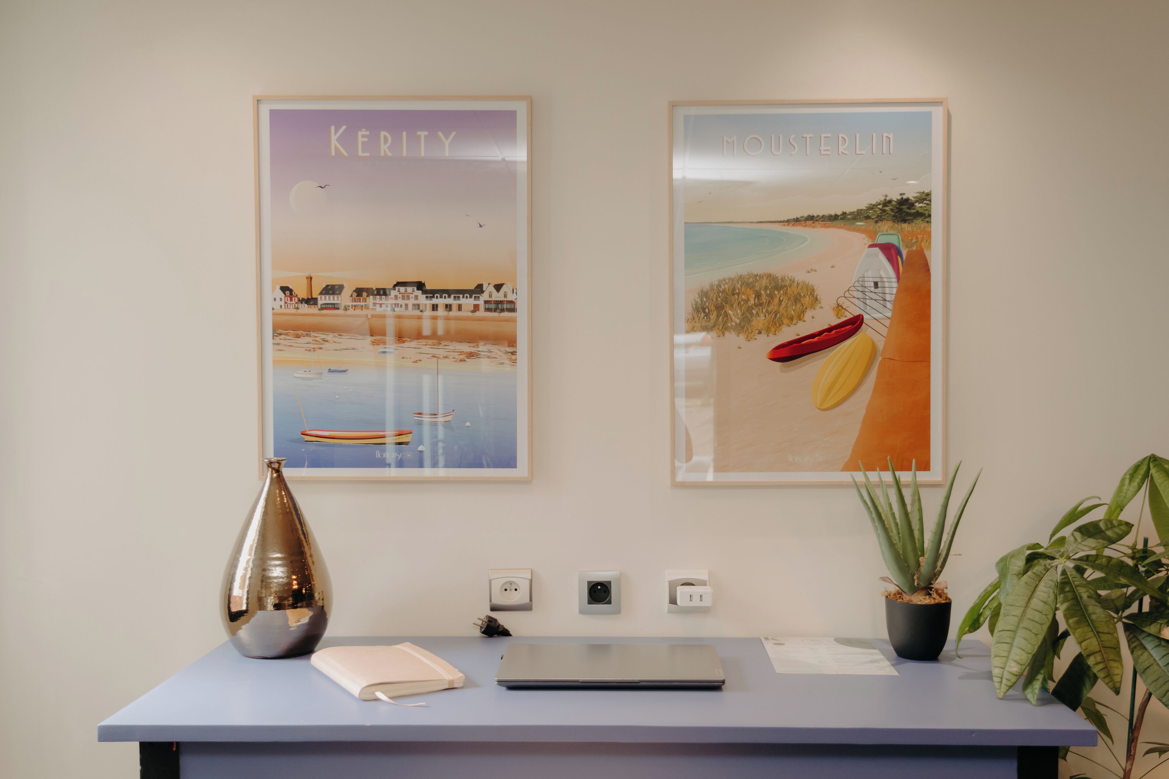 A reception desk with two framed posters, a laptop, a vase, and a potted plant.
