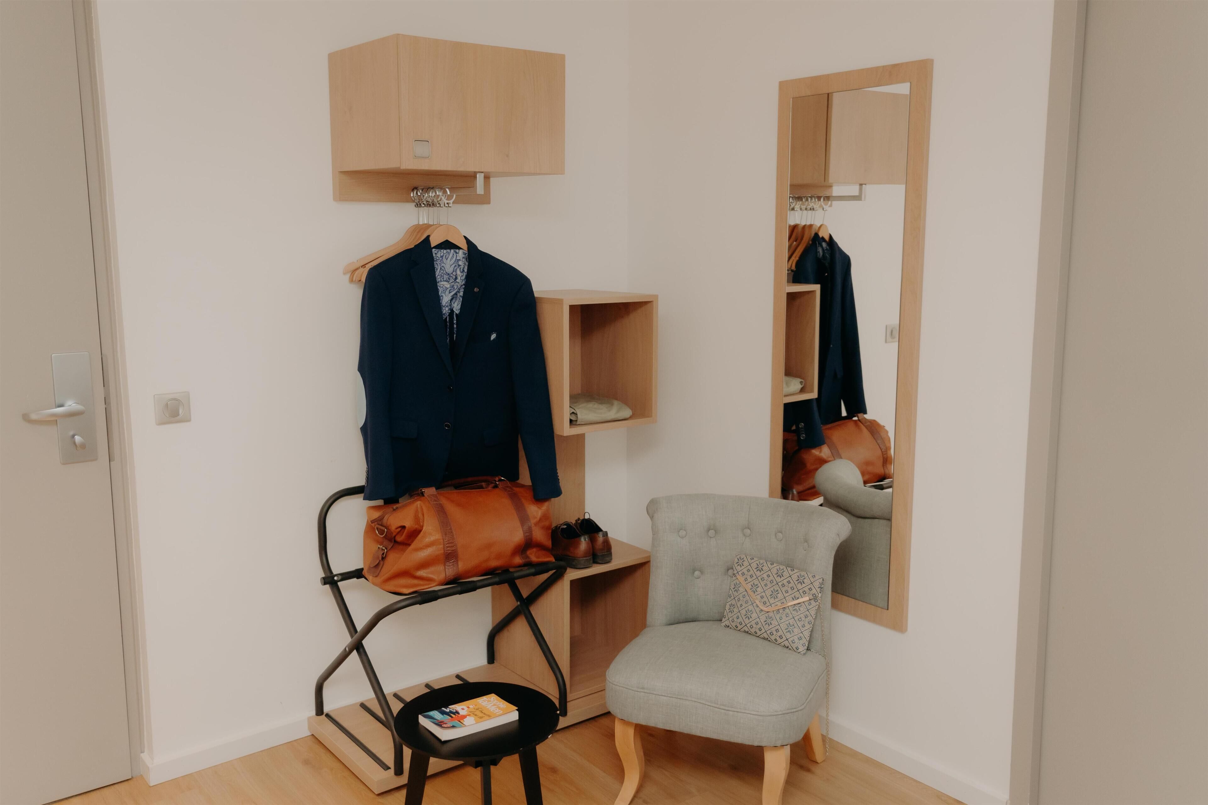 A room with a wooden wardrobe, a chair, a small table with a book, and a hanging bag.