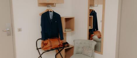 A room with a wooden wardrobe, a chair, a small table with a book, and a hanging bag.
