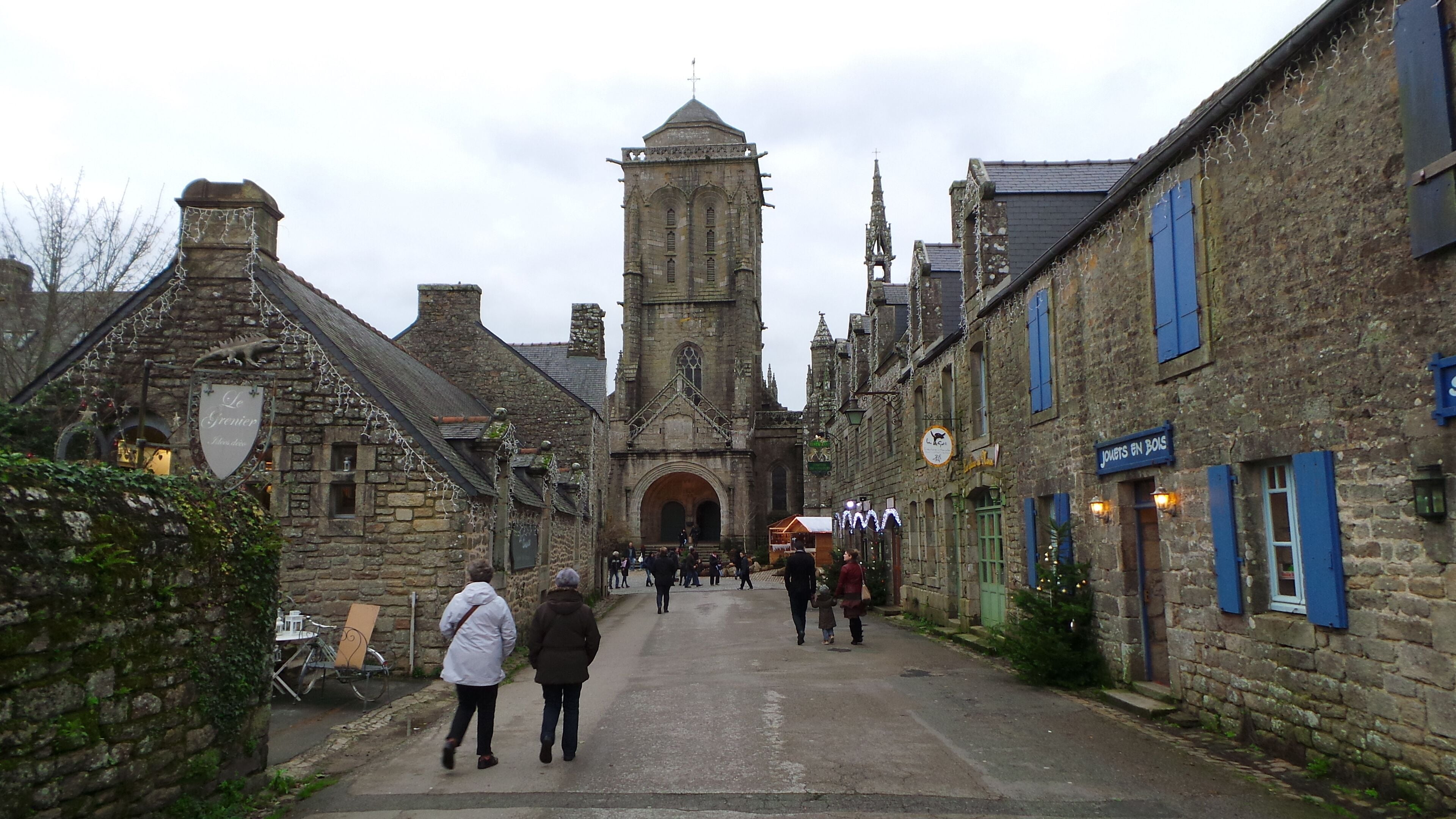 A narrow street with historic stone buildings, a prominent church tower, and a sign for 'Le Syrenier'.