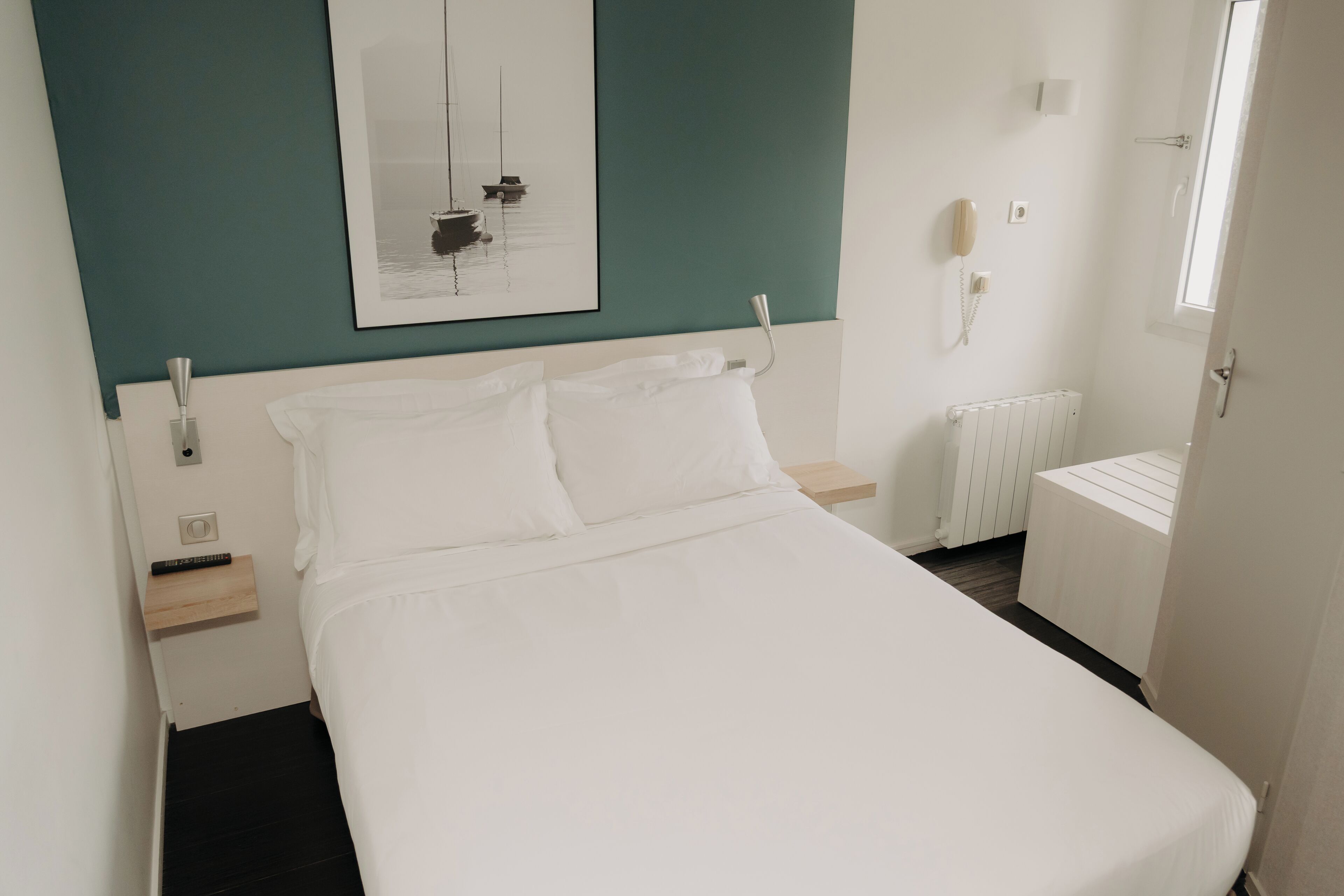 A neatly made bed in a hotel room with a teal accent wall, a white bedside table, a white radiator, and a telephone on the wall.