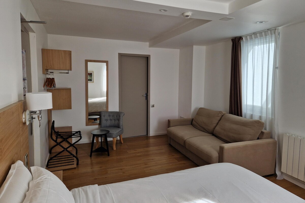 A hotel room with a bed, a sofa, a chair, a small table, a mirror, and a window with curtains.