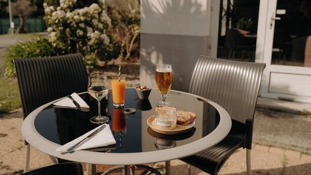 Una mesa redonda con una copa de vino blanco, un vaso de zumo de naranja, una cerveza, un plato con pan y un frasco de crema, y una servilleta.