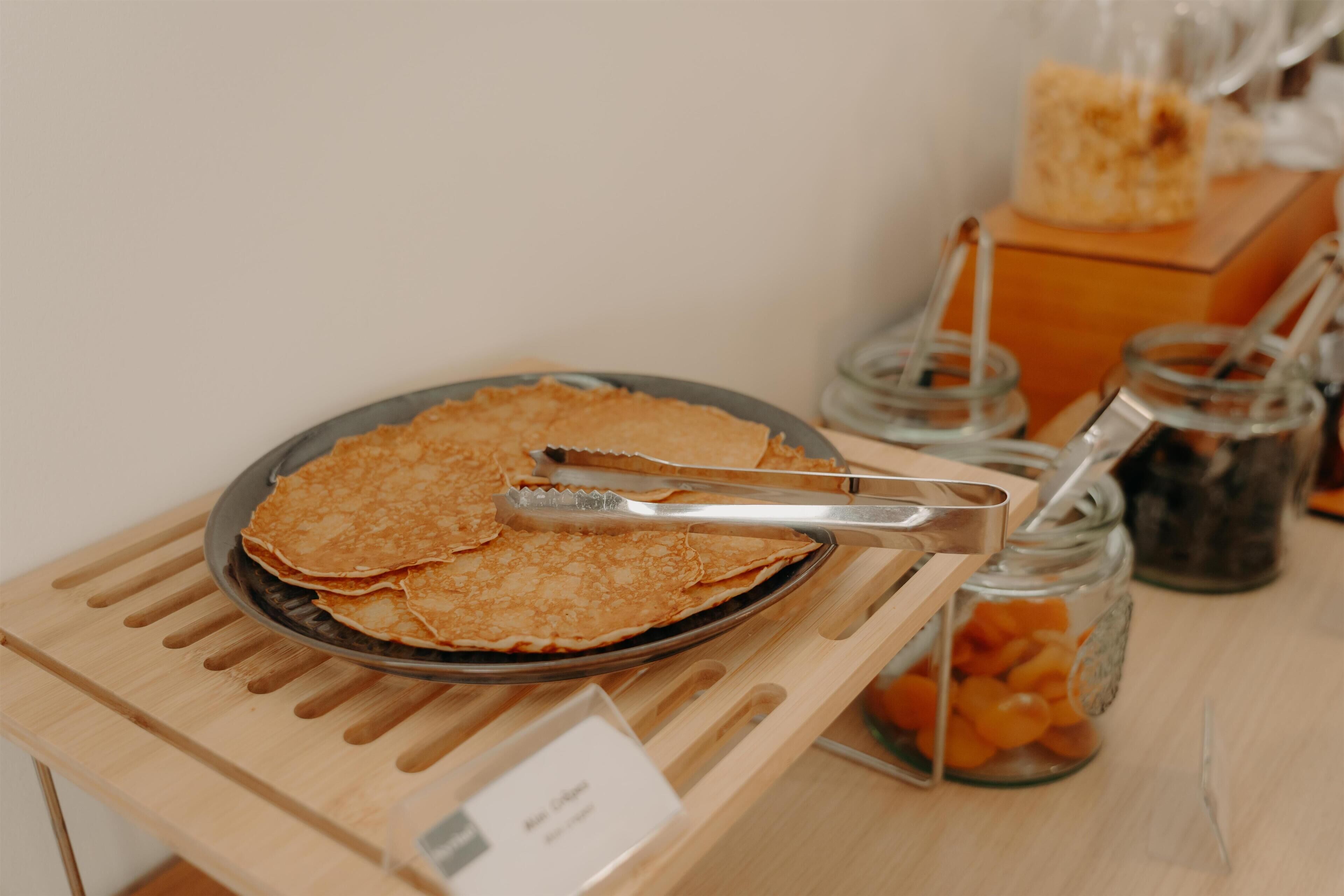 A tray of crepes on a wooden stand with tongs, a jar of orange slices, and a jar of blackberries.