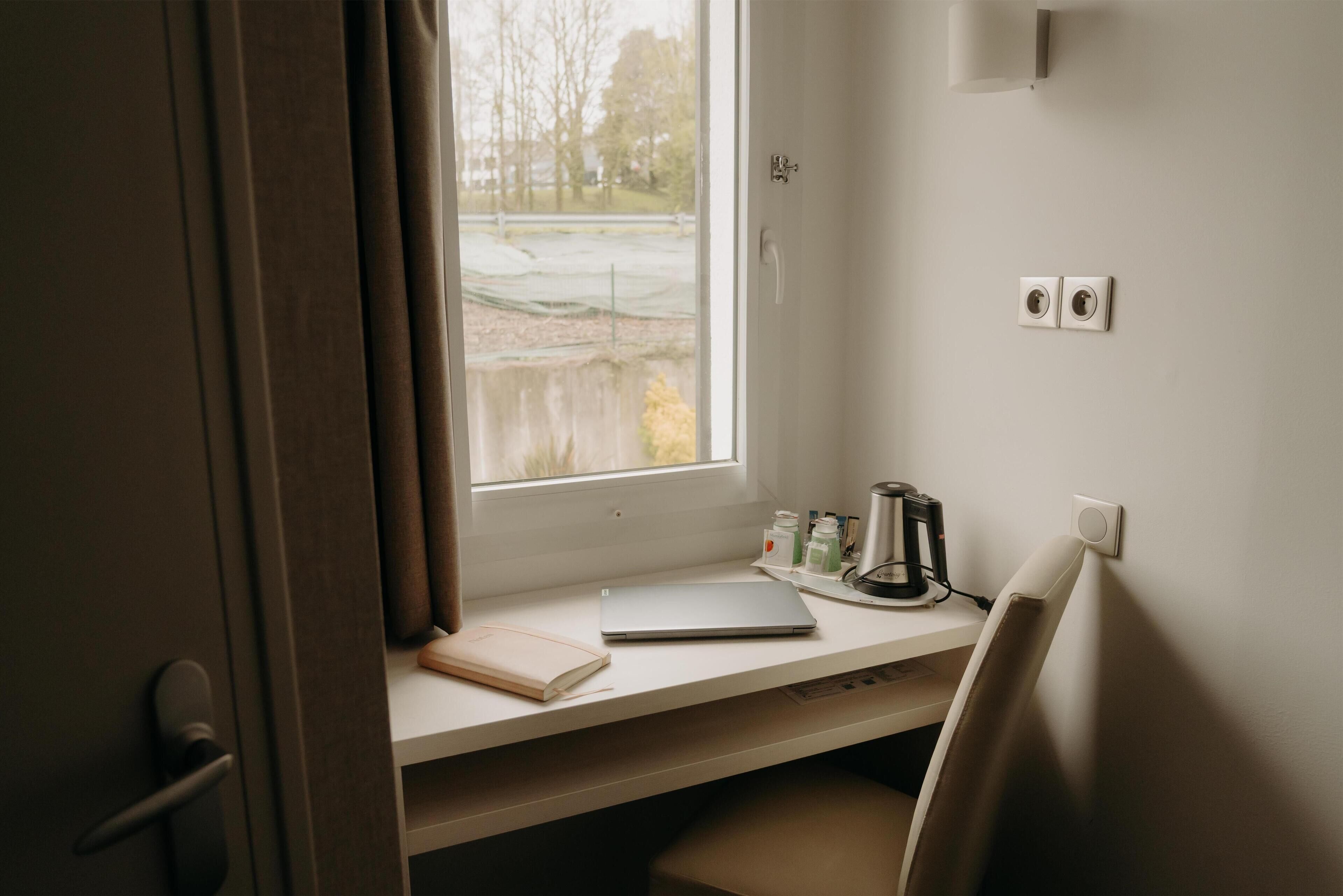 Una habitación con escritorio, laptop, cuaderno, hervidor y ventana con vistas a un parque.