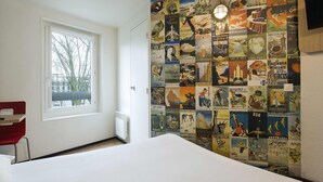 A hotel room with a large wall of vintage travel posters, a window with a view of trees, and a red chair. - hotelF1 Cergy (Cergy)