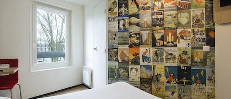 Habitación de hotel con una gran pared de carteles publicitarios de viajes antiguos, una ventana con vistas a árboles y una silla roja.