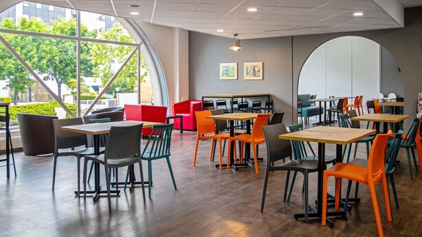 A modern cafe with wooden tables and chairs, large windows, and a curved wall with artwork. - hotelF1 Cergy (Cergy)
