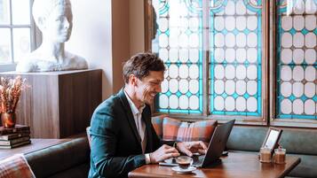 A man working on a laptop at a wooden table with a glass of coffee, in a room with a bust sculpture and patterned windows.